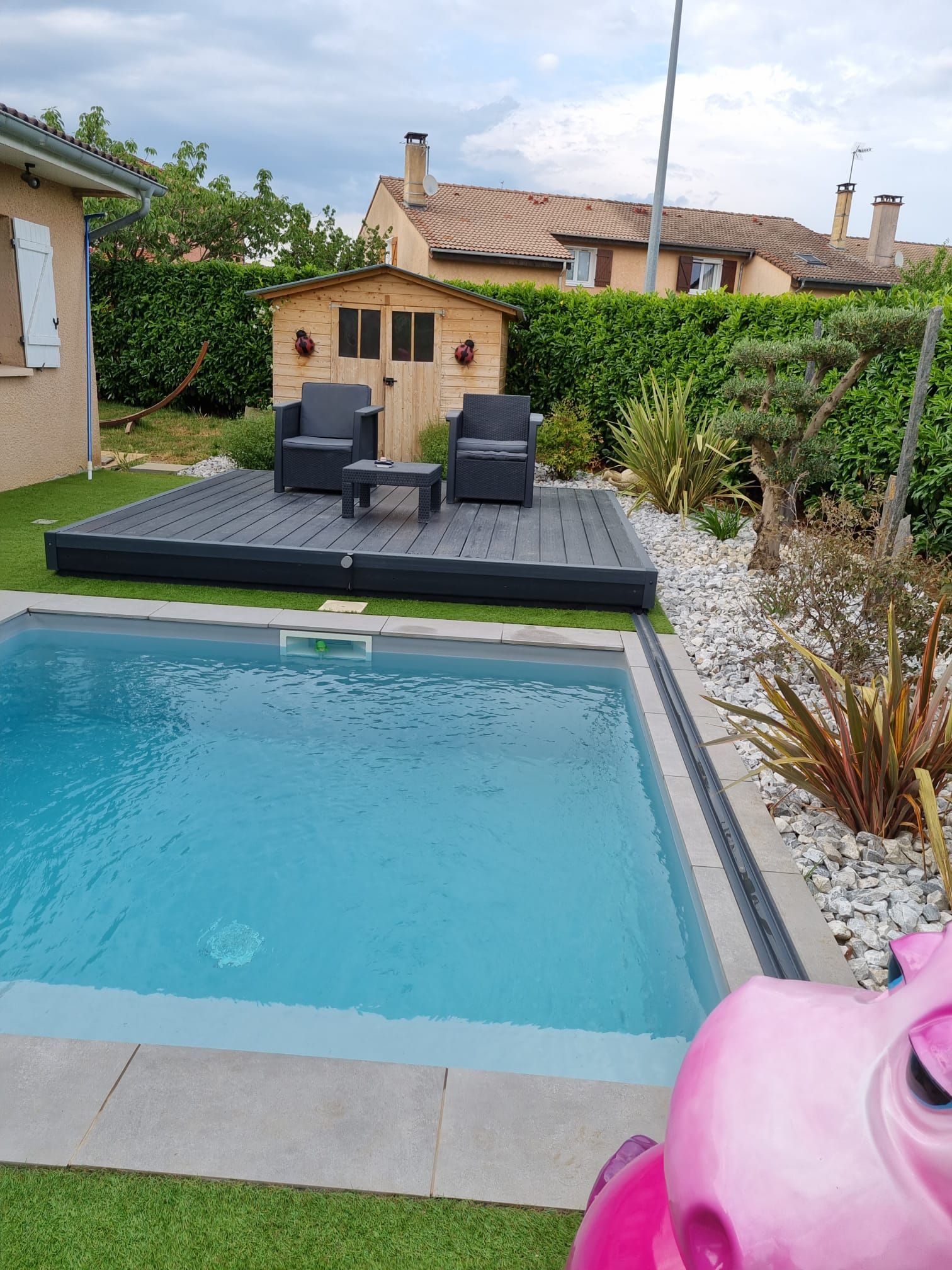 Piscine dans la cour arrière avec chaises longues, petit abri de jardin et haies sous un ciel nuageux