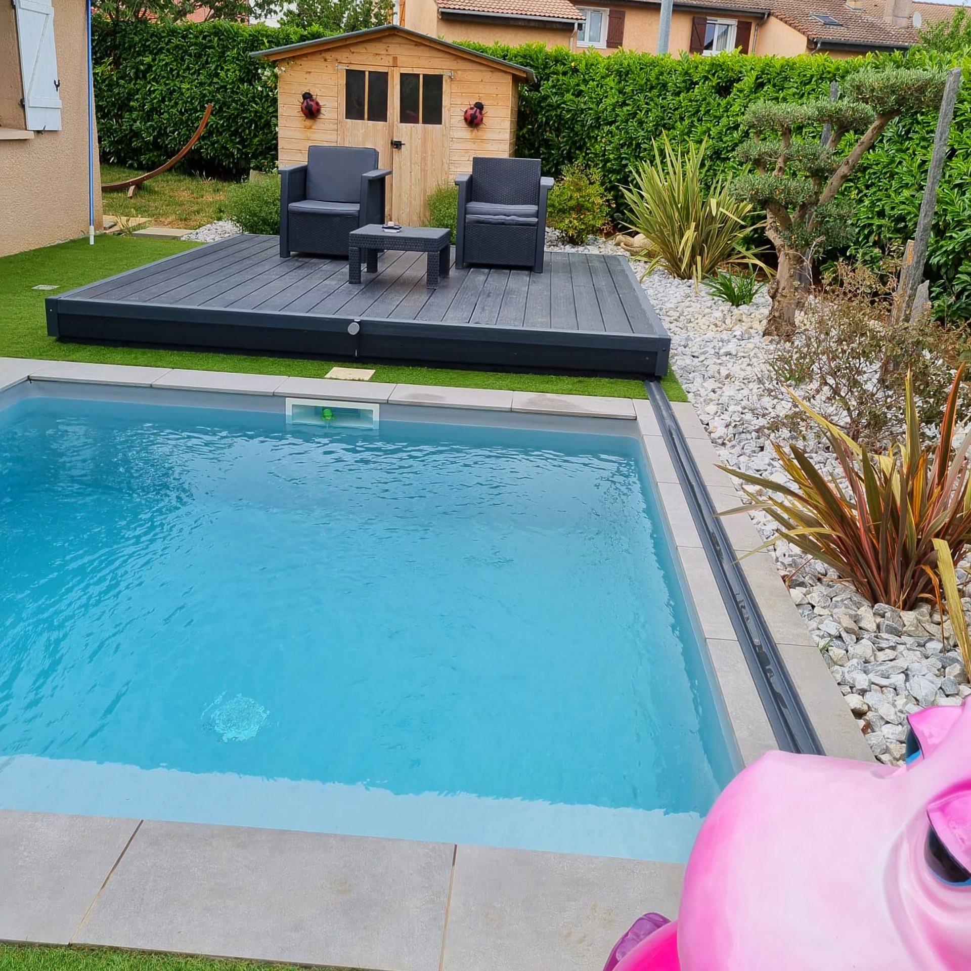 Piscine dans le jardin, à côté d'une terrasse surélevée avec chaises de jardin et un petit abri de jardin, le tout entouré de verdure.
