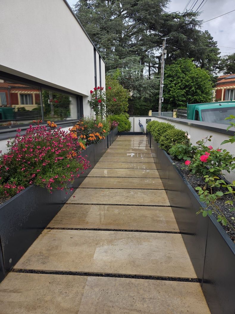 Allée de jardin longeant un bâtiment blanc, bordée de fleurs colorées et de verdure après la pluie.