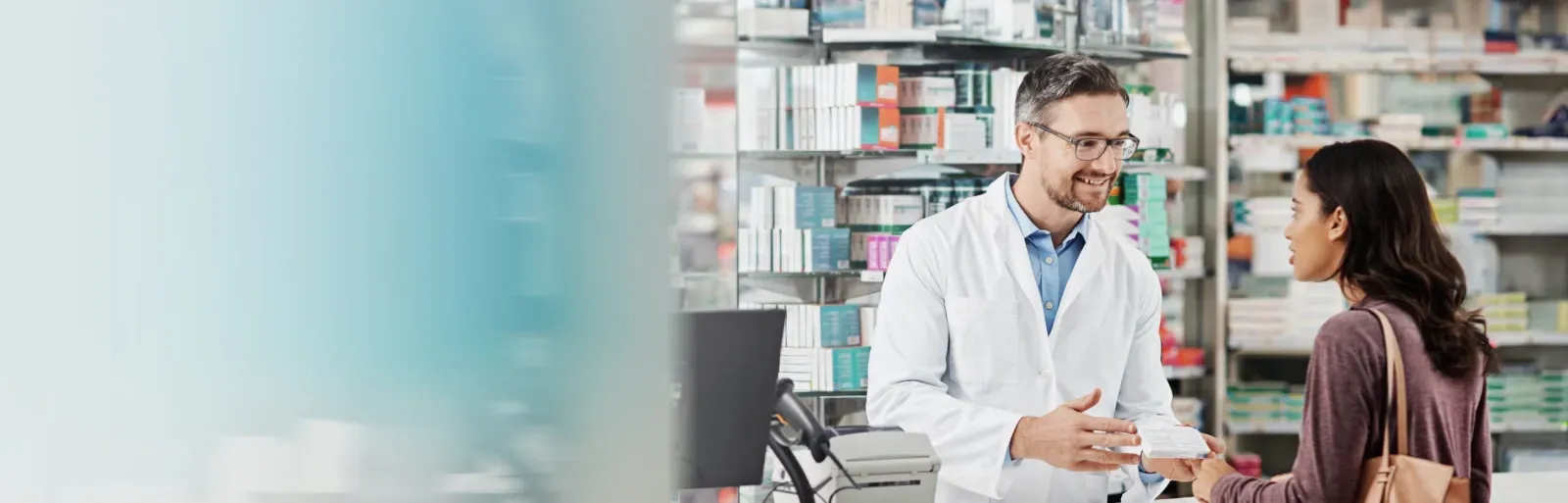 Ein Apotheker unterhält sich mit einem Kunden an einem Apothekenschalter; im Hintergrund sind Regale mit Medikamenten zu sehen.