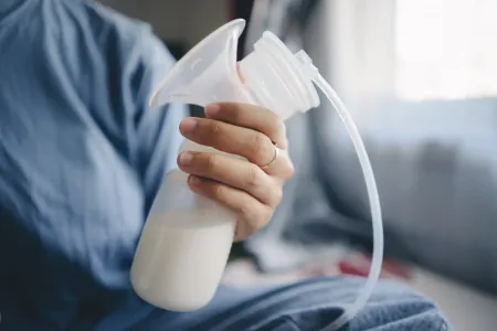 Eine Hand hält neben einem Bett eine weiße Milchpumpenflasche, in der sich Milch befindet und an der ein Schlauch befestigt ist.