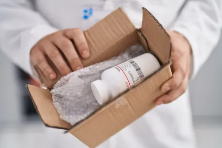 Hände öffnen einen Karton mit Luftpolsterfolie und einer weißen Medikamentenflasche darin.