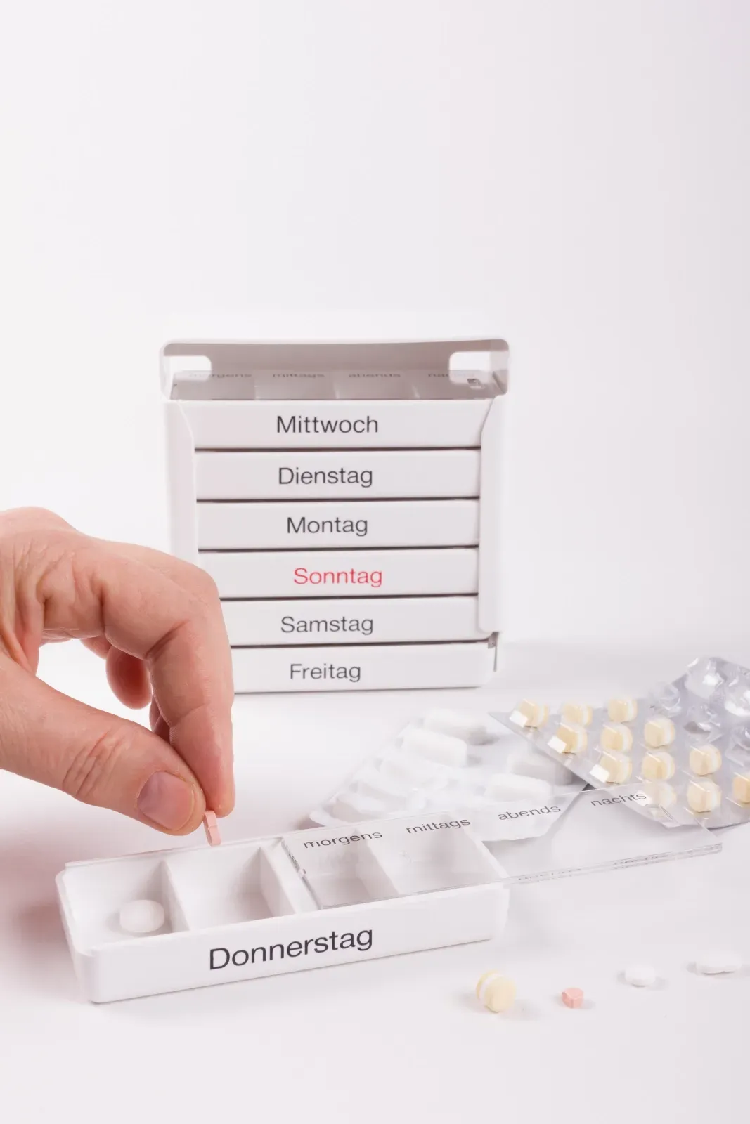 Eine Hand legt Tabletten in einen beschrifteten Wochenpillenorganizer neben einer Blisterpackung.