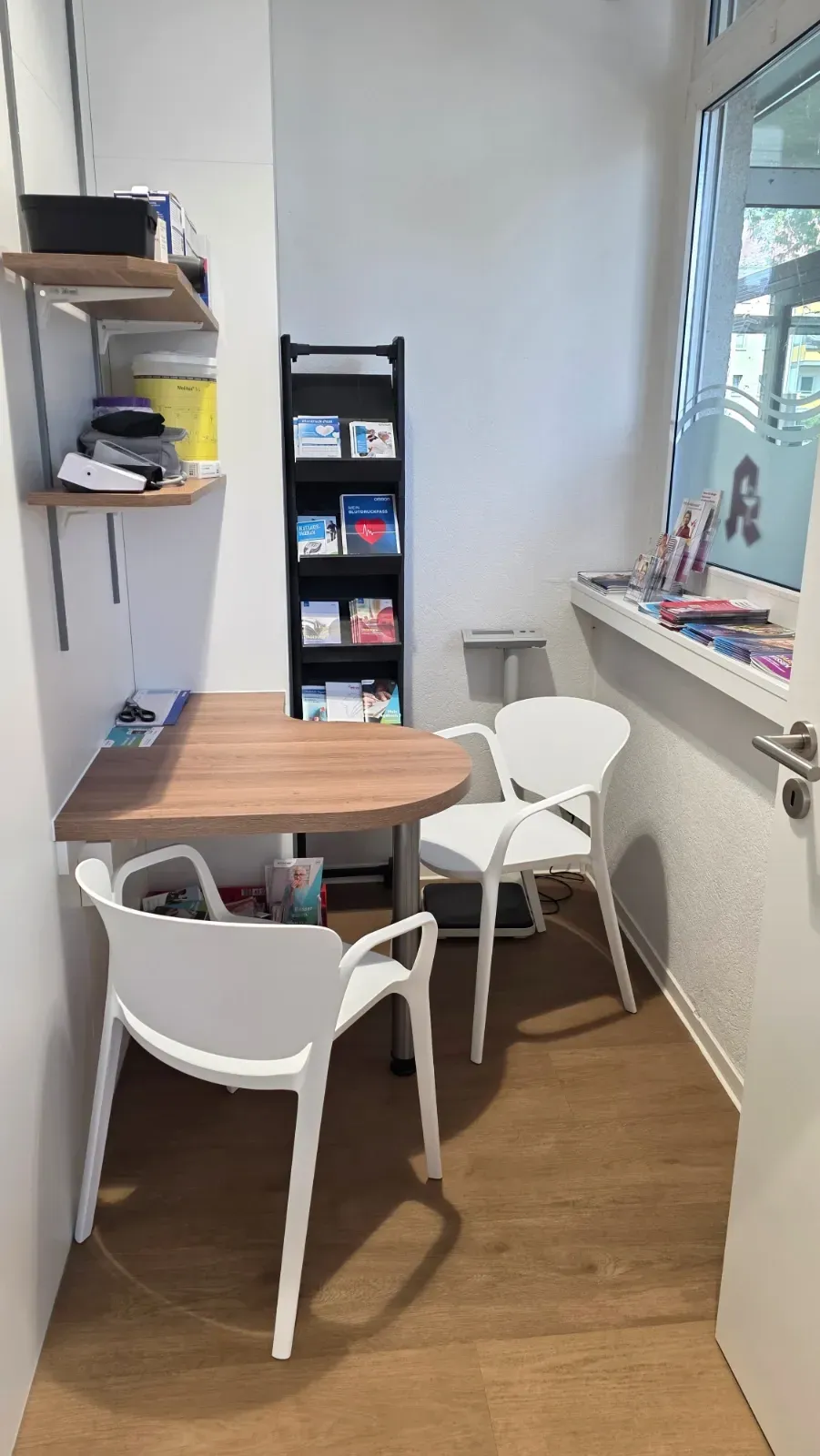 Kleine Büroecke mit einem runden Tisch, drei weißen Stühlen und einem an der Wand befestigten Prospektständer.
