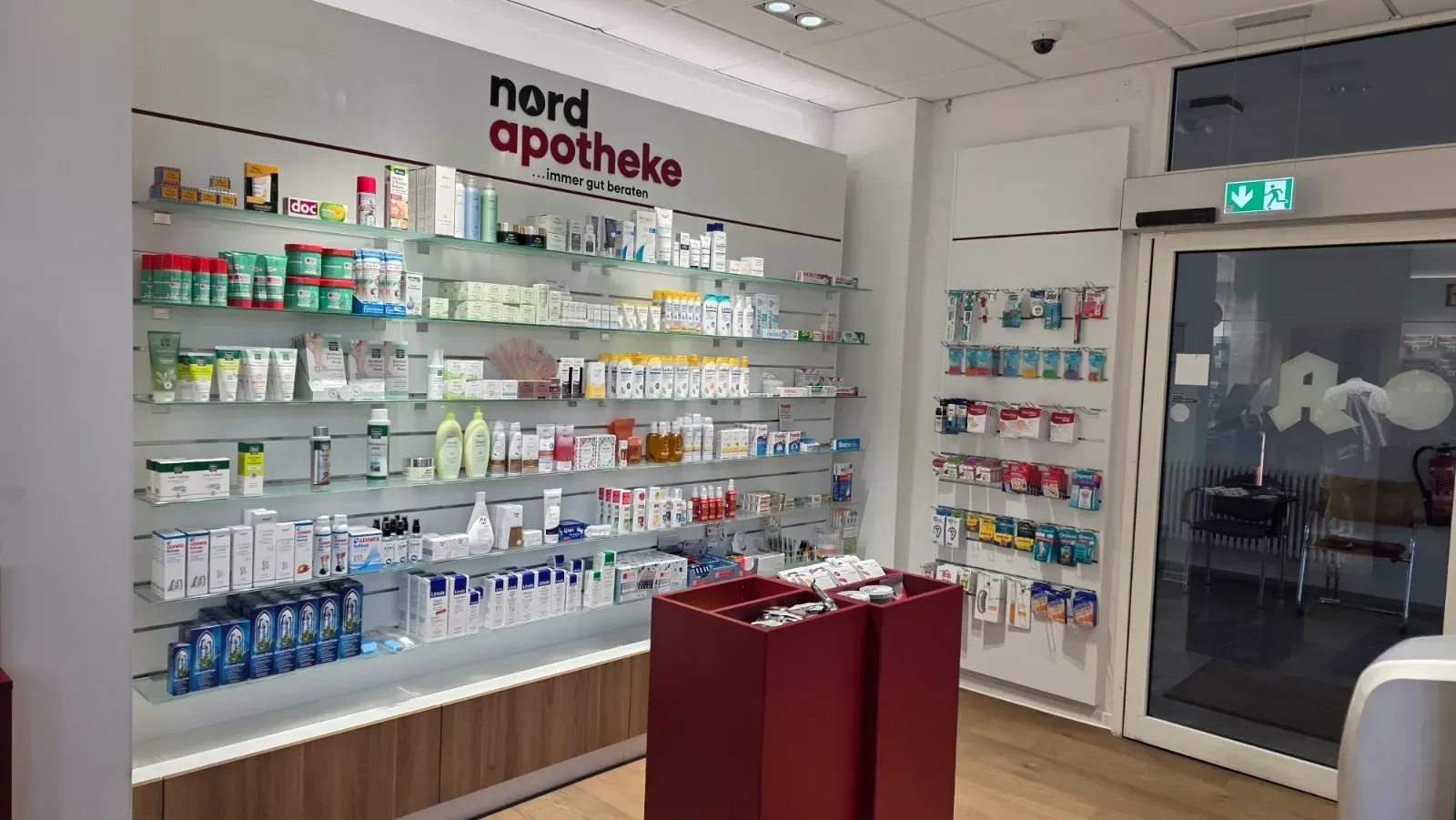 Apothekeninterieur mit Wandregalen voller Produkte, rotem Präsentationstisch und gläserner Eingangstür