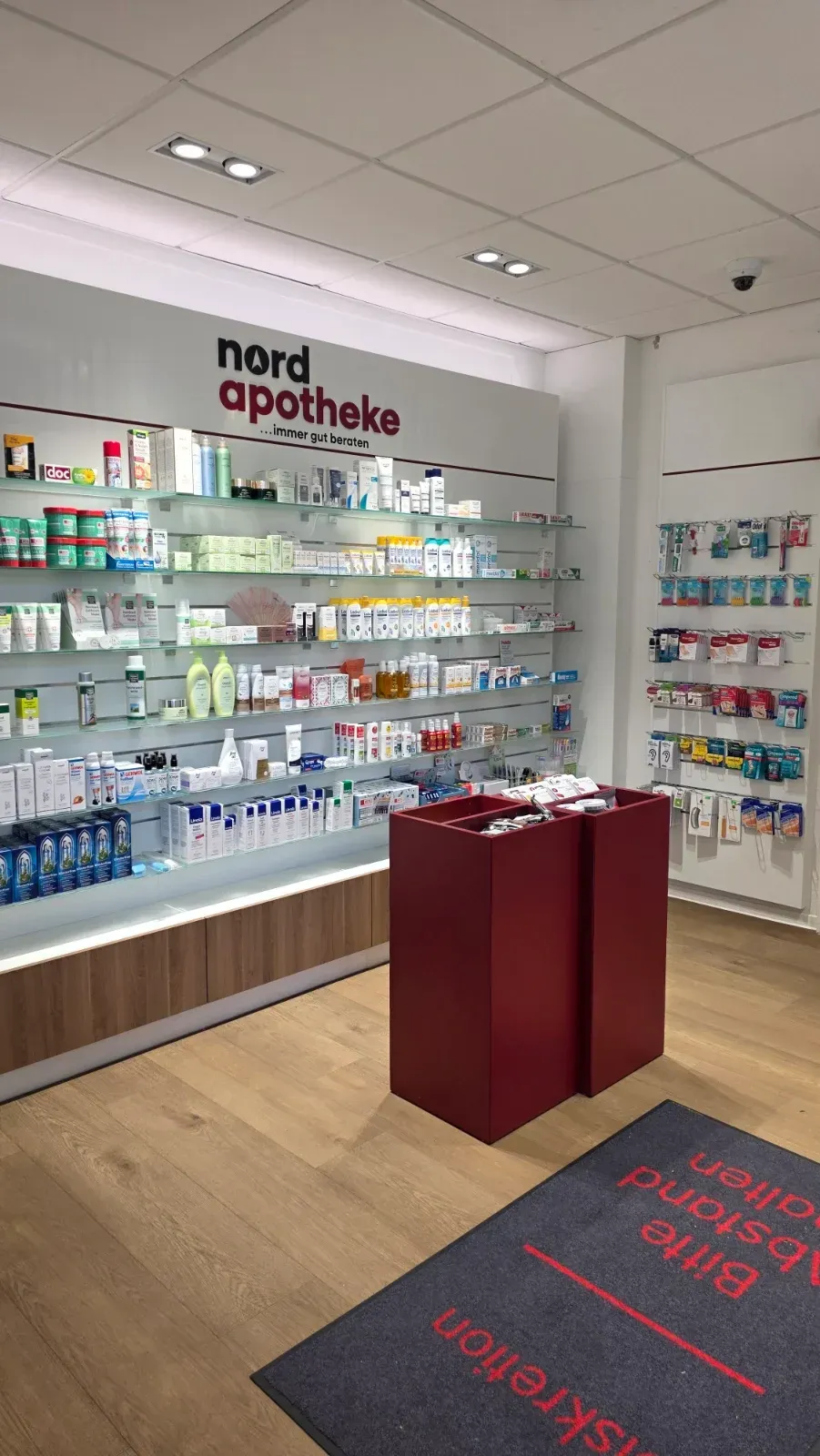 Apothekeninterieur mit Regalen voller Produkte, rotem Verkaufstresen und dem Schild „Apotheke“ an der Wand.