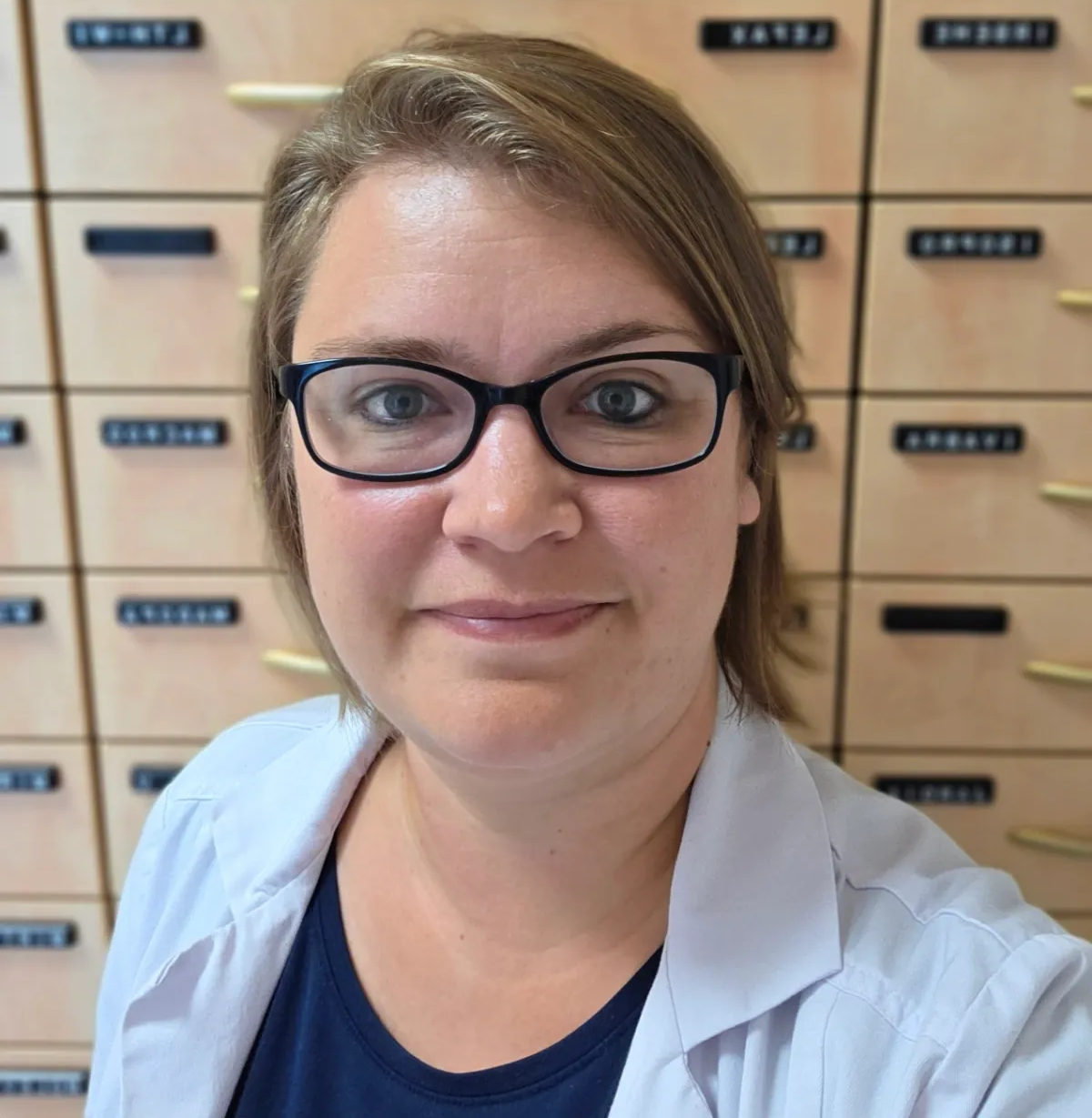Eine lächelnde Person mit Brille und weißem Kittel vor beschrifteten Lagerregalen.