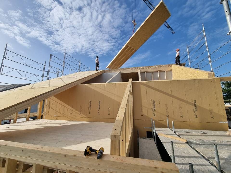 Deux ouvriers se tiennent debout sur la charpente en bois d'un bâtiment en construction tandis qu'une grue abaisse un grand panneau de toiture en bois.