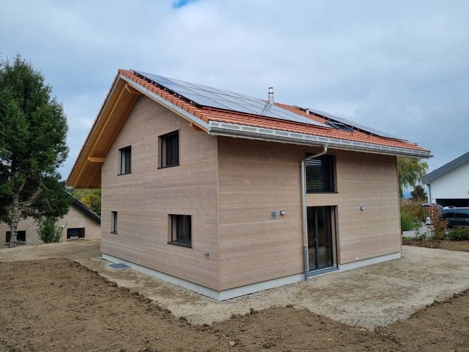 Une maison en bois neuve, avec des panneaux solaires sur un toit de tuiles, entourée de terre nue par temps nuageux.