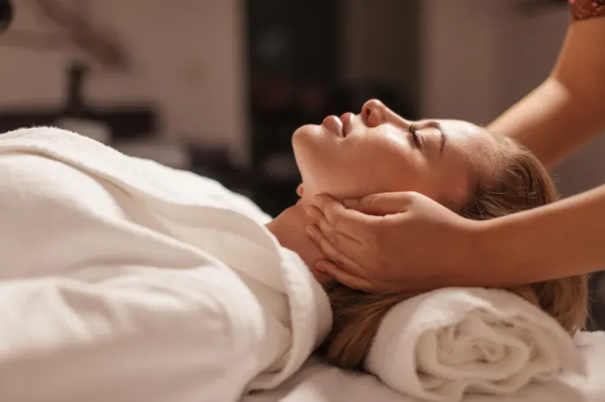Persona recibiendo un masaje facial mientras está tumbada en una camilla de spa con una toalla blanca sobre la cabeza.