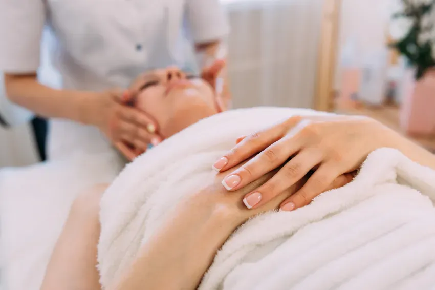 Persona recibiendo un masaje facial con toallas blancas y una mano apoyada en la frente.