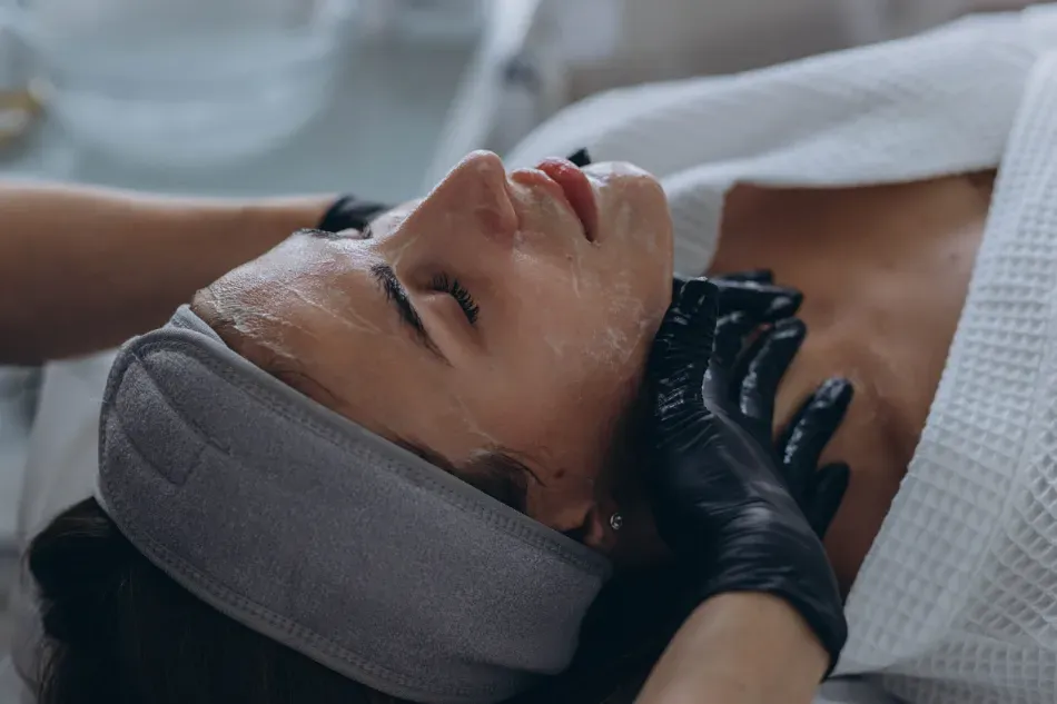 Persona recibiendo un tratamiento facial mientras está tumbada en una camilla de spa, con las manos enguantadas de negro sobre la cara.