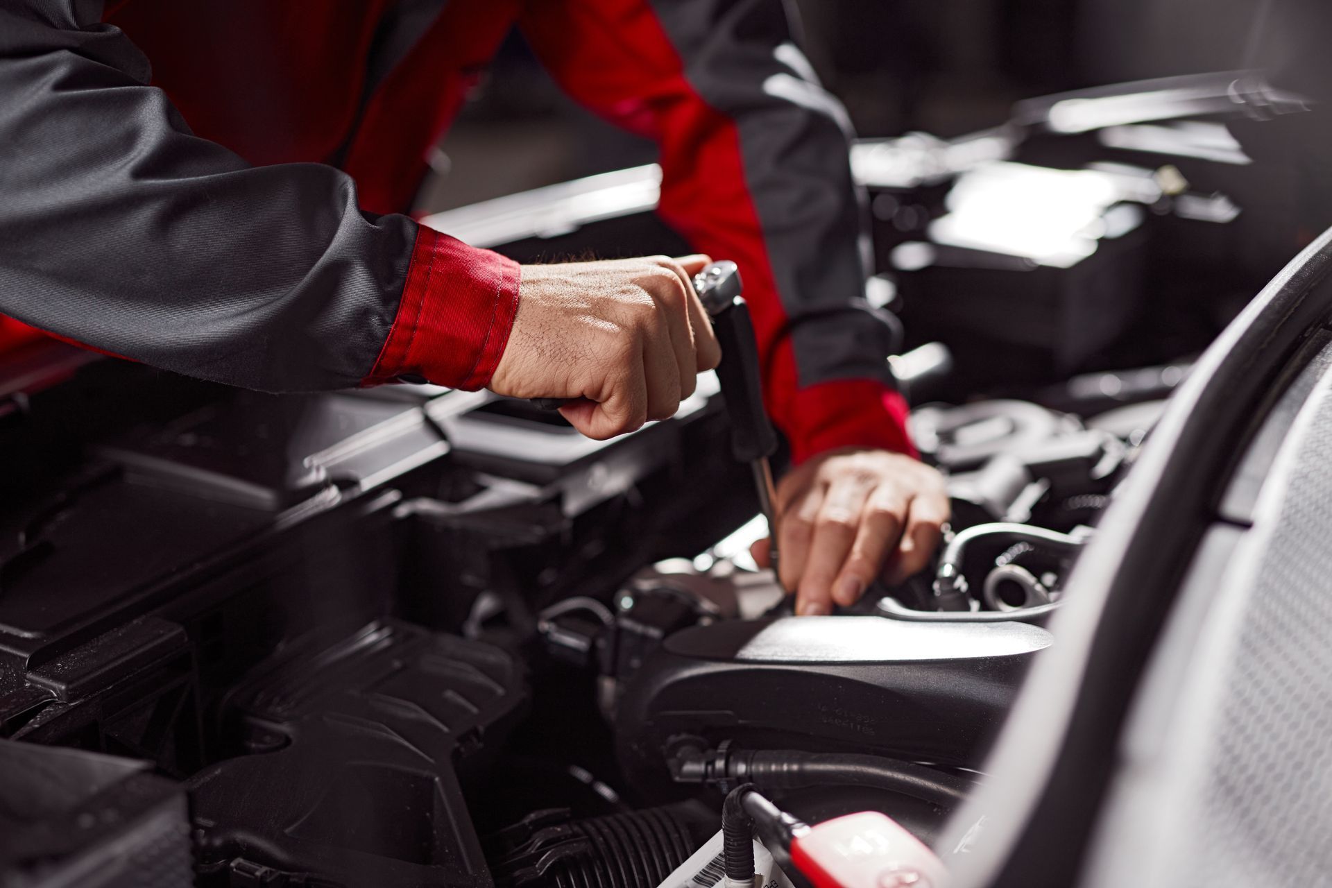 Mécanicien travaillant sur un moteur de voiture avec une clé, portant un uniforme rouge et gris dans un atelier.