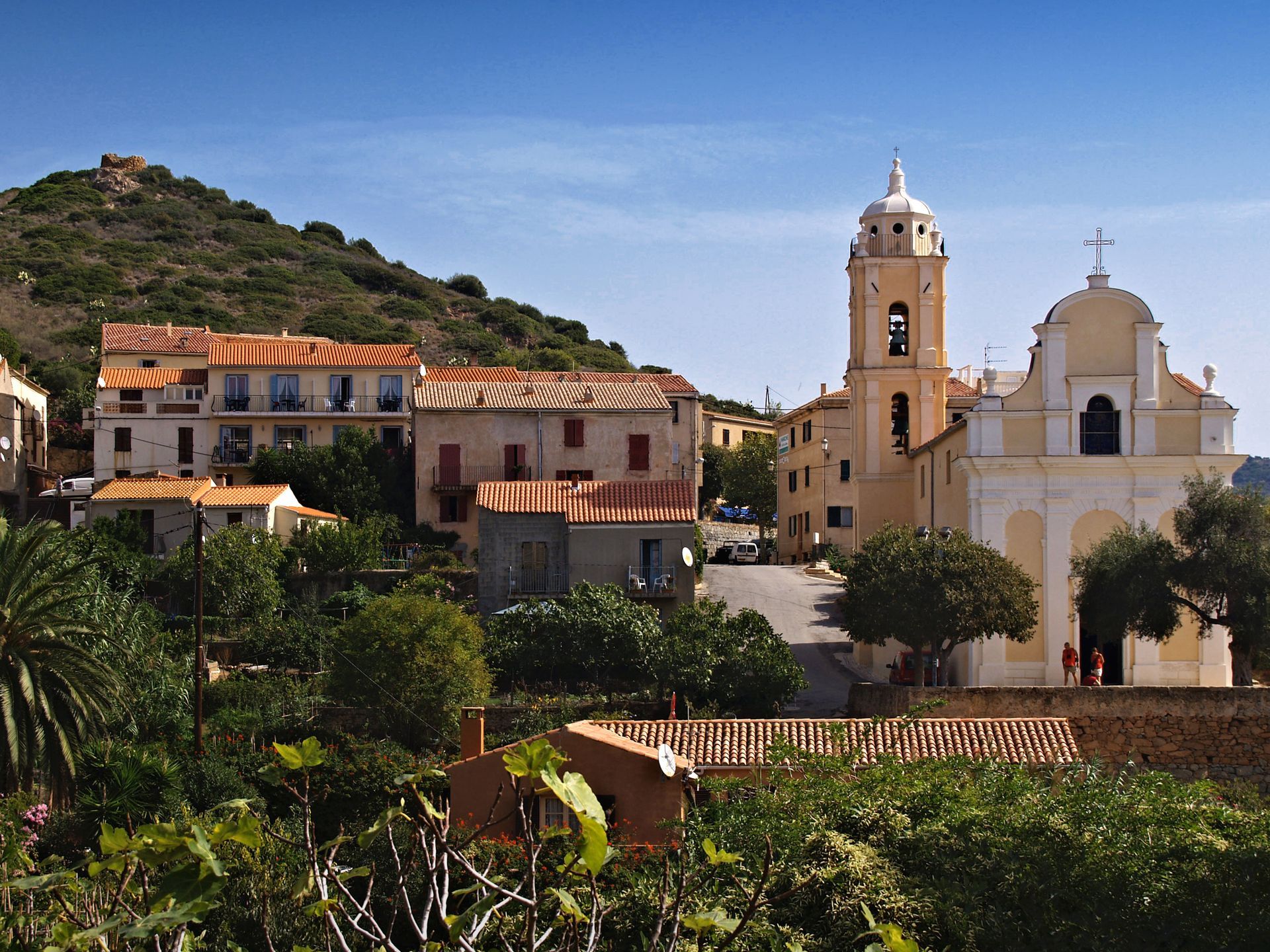 Village avec église et clocher contre une montagne, sous un ciel bleu clair.