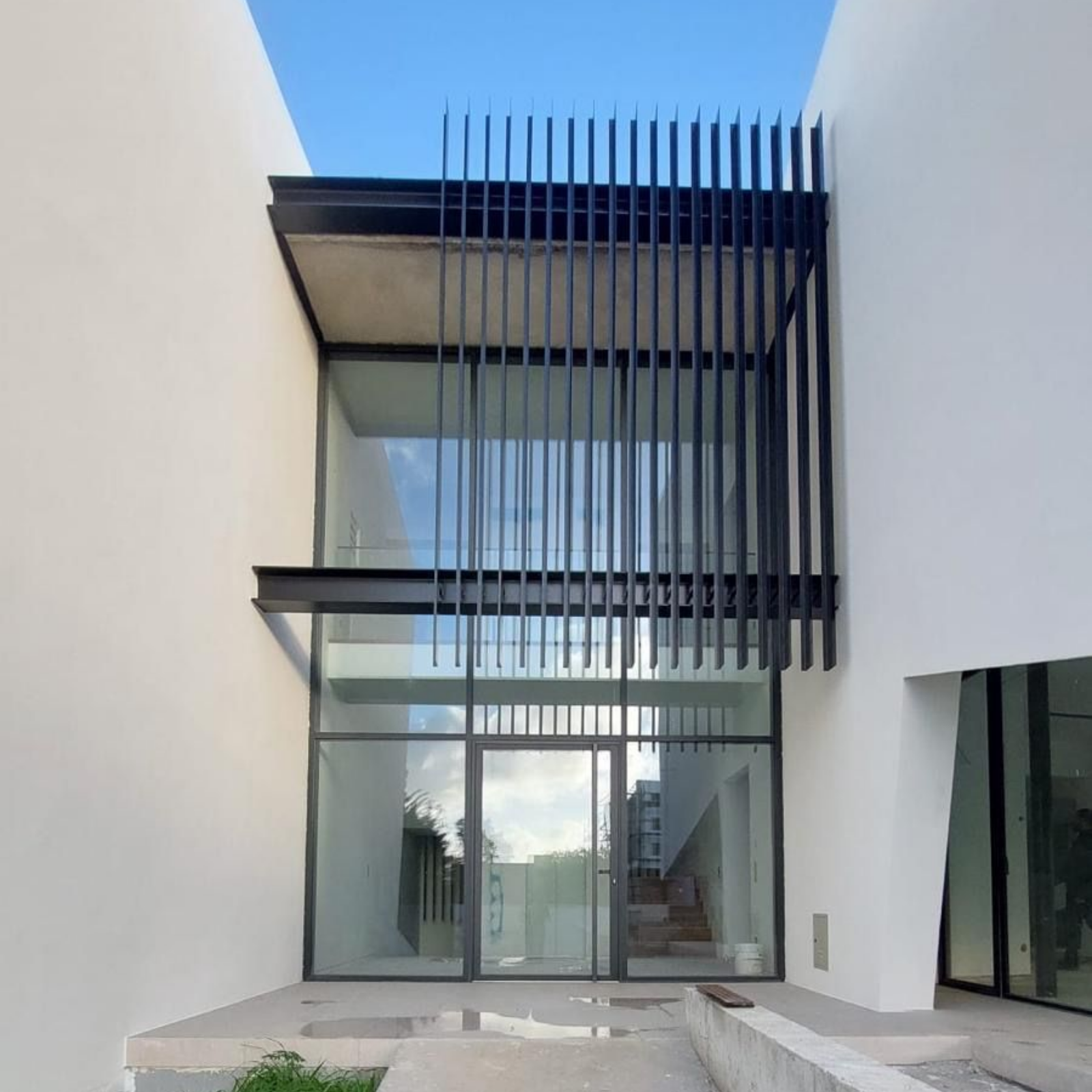 Façade moderne blanche avec de grandes baies vitrées, des lamelles verticales noires et une petite cour en béton.