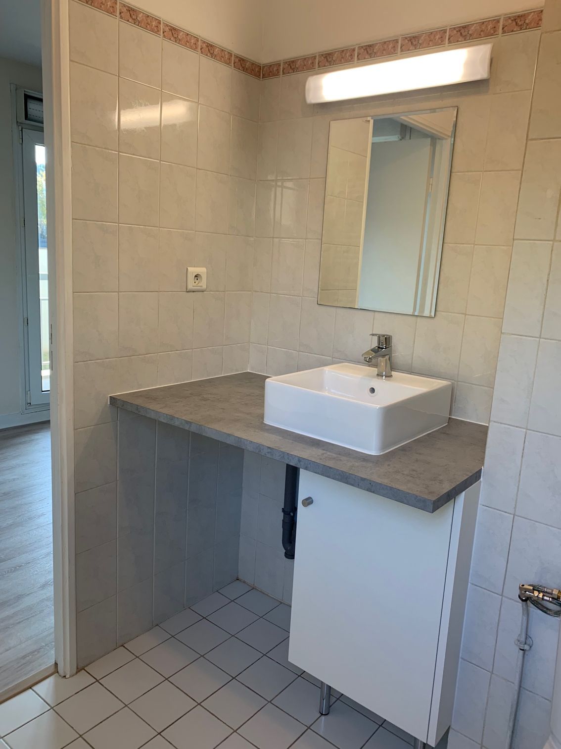 Meuble-lavabo de salle de bain avec lavabo blanc, comptoir gris, miroir et murs et sol carrelés.