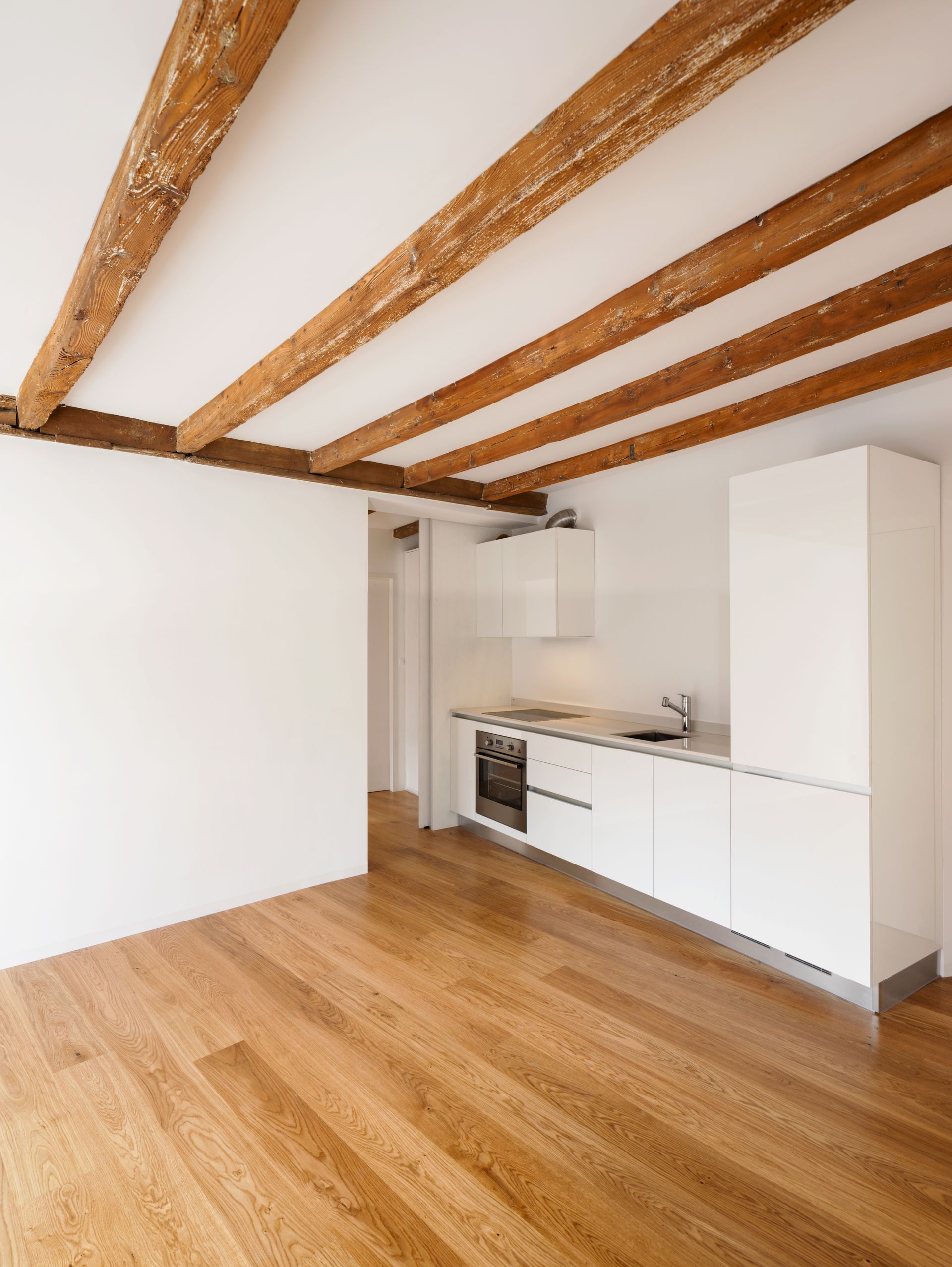 Pièce lumineuse et vide avec poutres apparentes, parquet clair et petite cuisine blanche