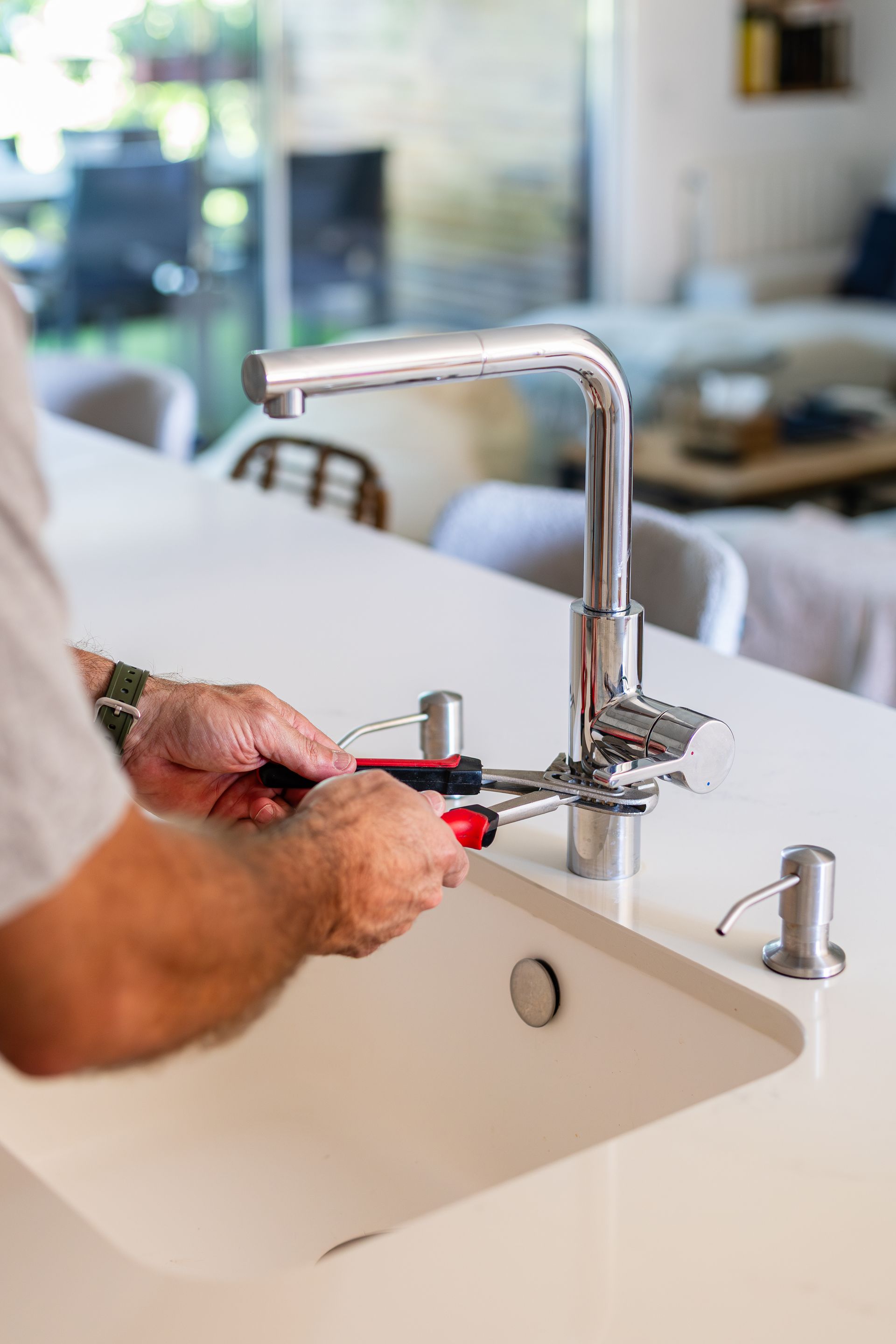 Une personne utilise une clé à poignée rouge pour serrer un robinet de cuisine dans un évier blanc.