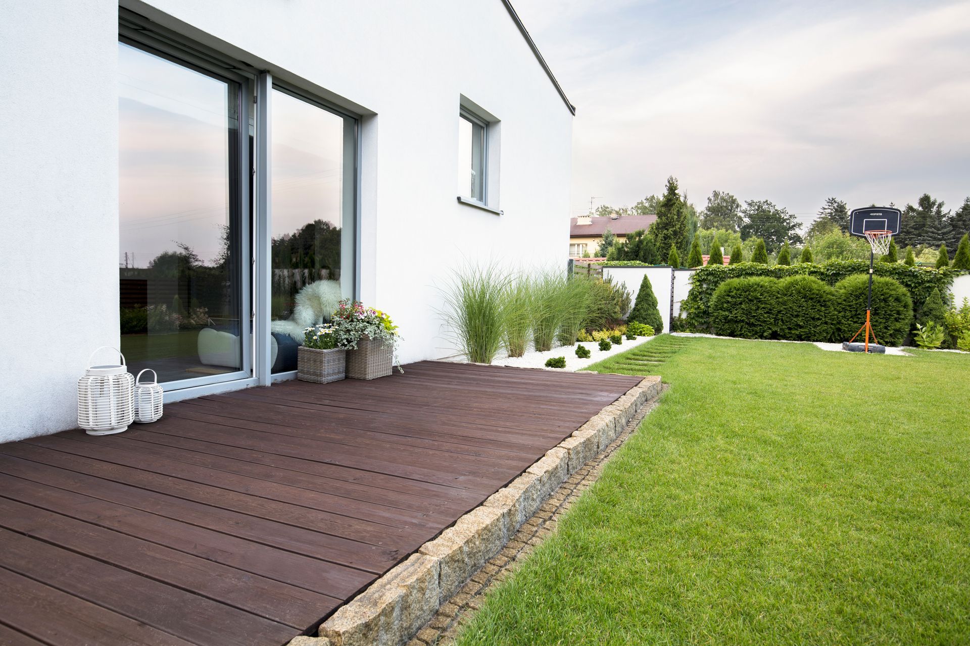 Patio de maison moderne avec terrasse en bois, portes vitrées, plantes en pot et pelouse verte bordée d'une haie.