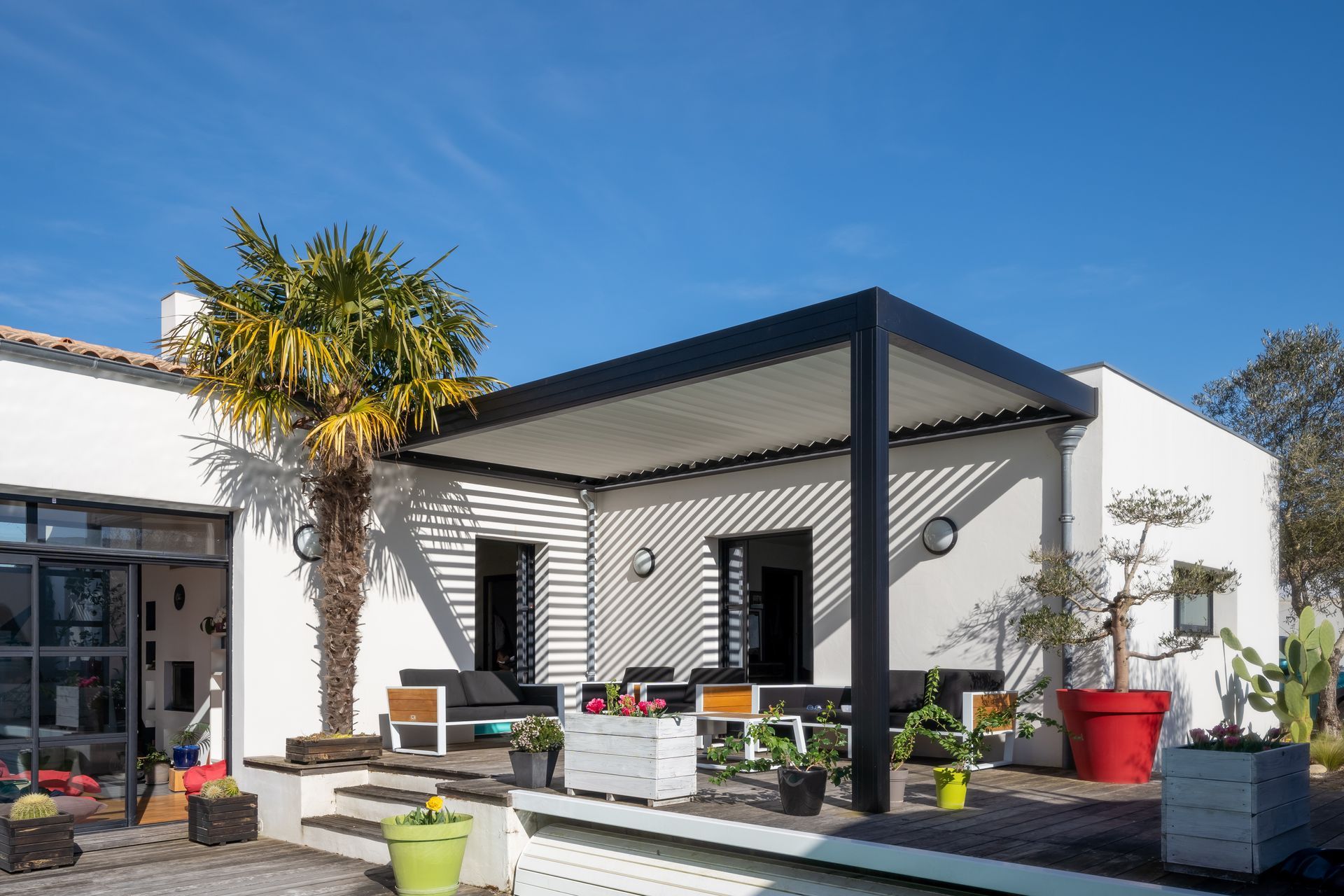 Patio d'une maison blanche moderne avec pergola noire, plantes en pot et sièges extérieurs sous un ciel bleu