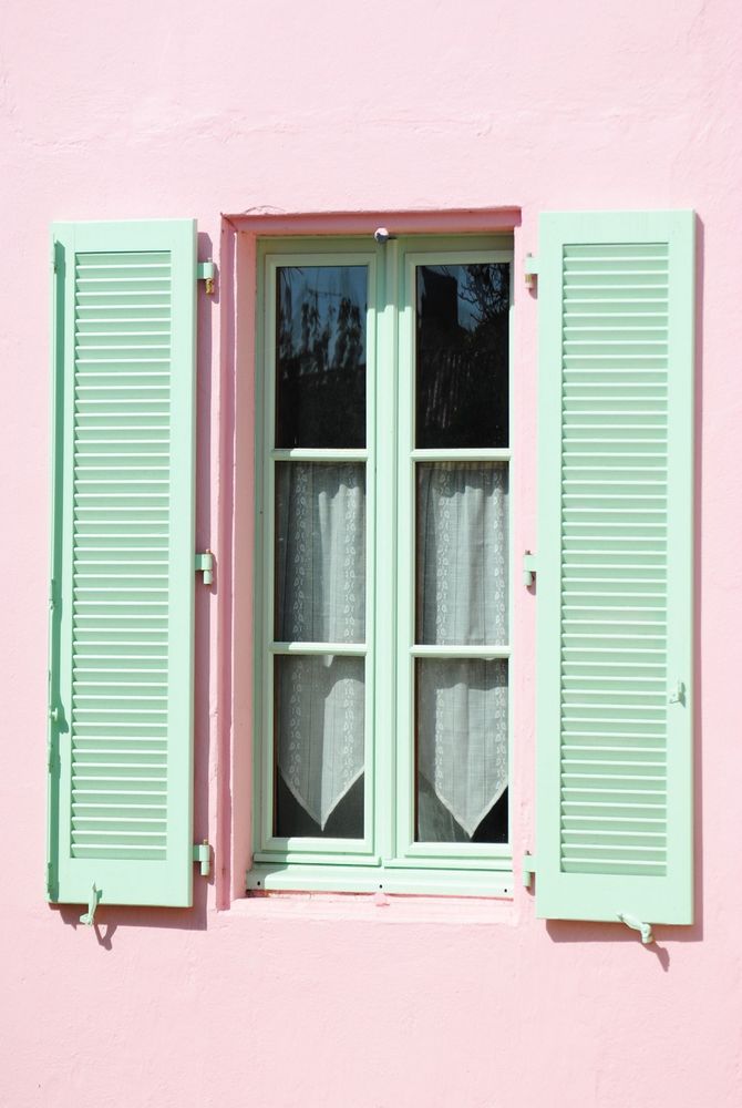 Mur rose pastel avec une fenêtre blanche encadrée de volets vert menthe.