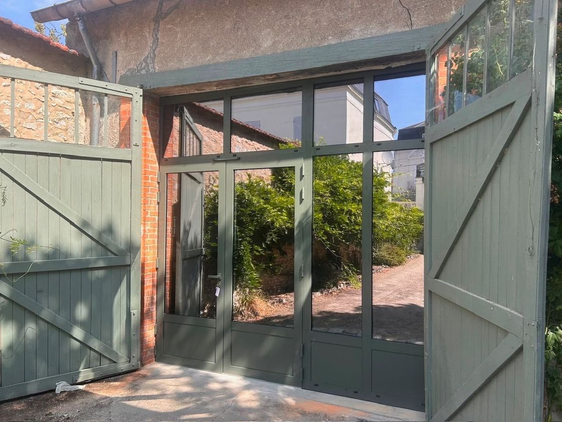 Portail de jardin ouvert avec panneaux de verre et chemin de briques menant à la verdure sous une entrée couverte