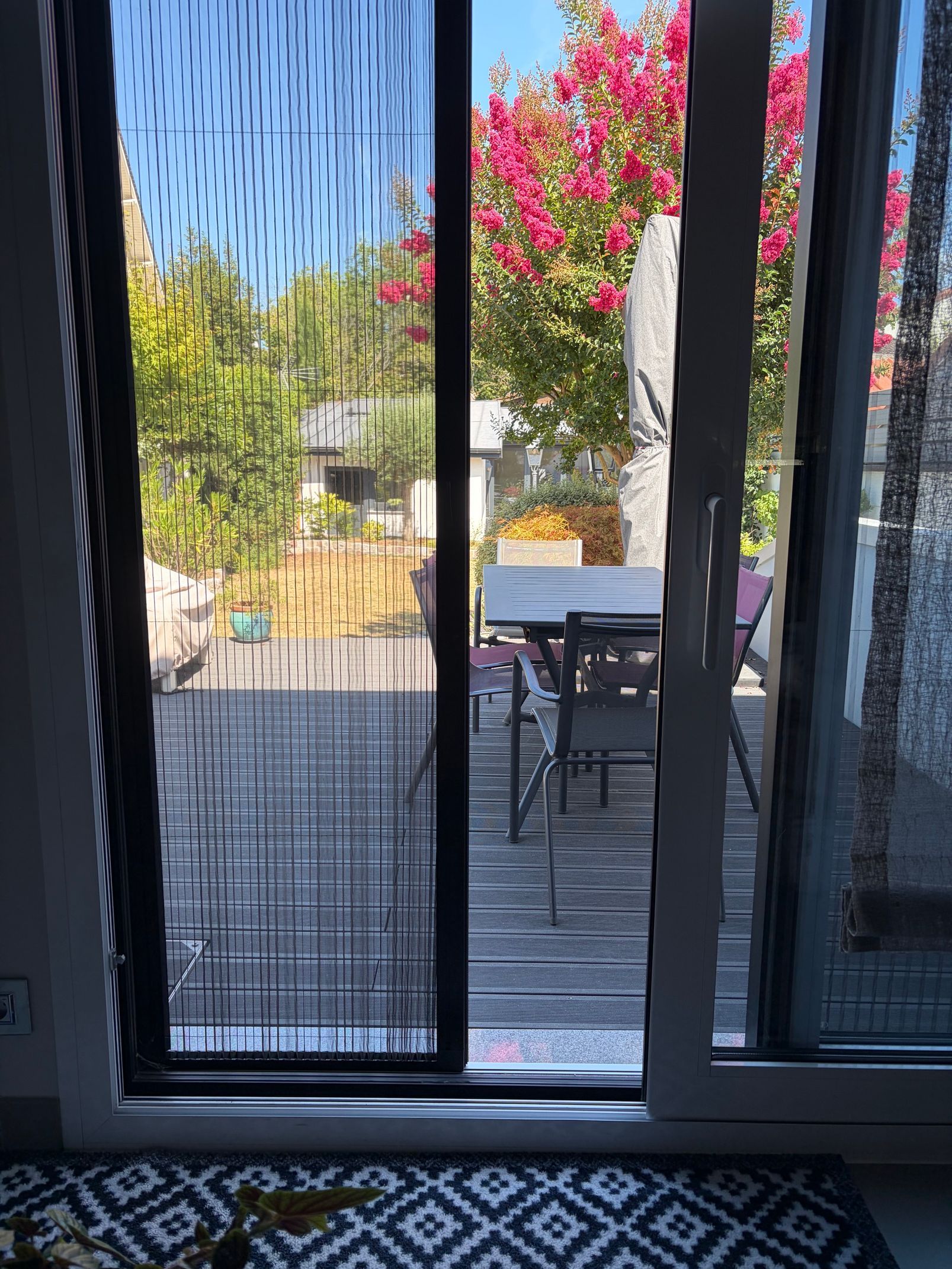 Porte coulissante donnant sur une terrasse ensoleillée avec table, chaises et un arbre à fleurs roses