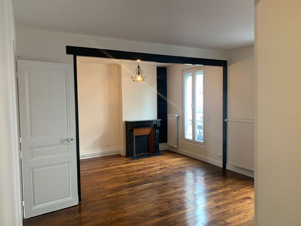 Pièce vide avec parquet, murs blancs, cheminée noire et porte vitrée.