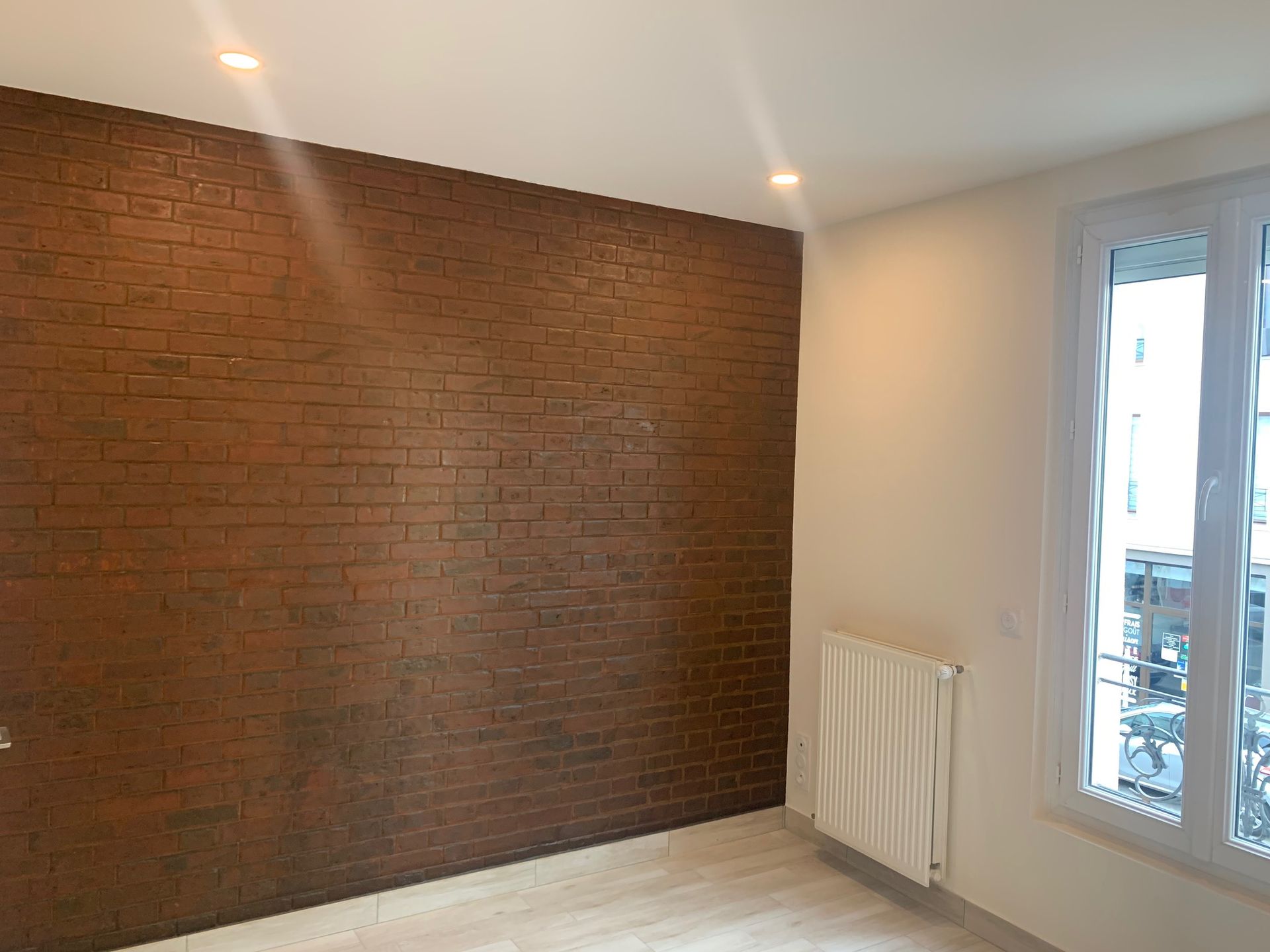 Pièce vide avec un mur en briques rouges, des murs blancs, un radiateur et une porte vitrée donnant sur un balcon.