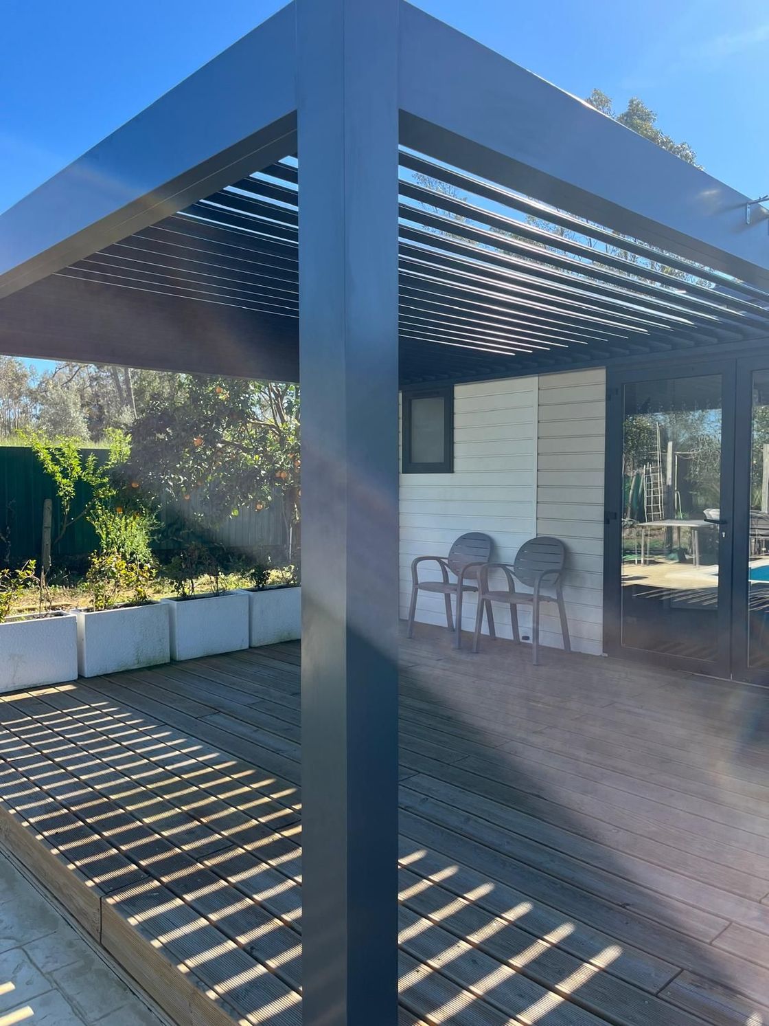Pergola de patio moderne avec toit à lattes projetant des ombres rayées sur un petit coin salon près d'un abri de jardin