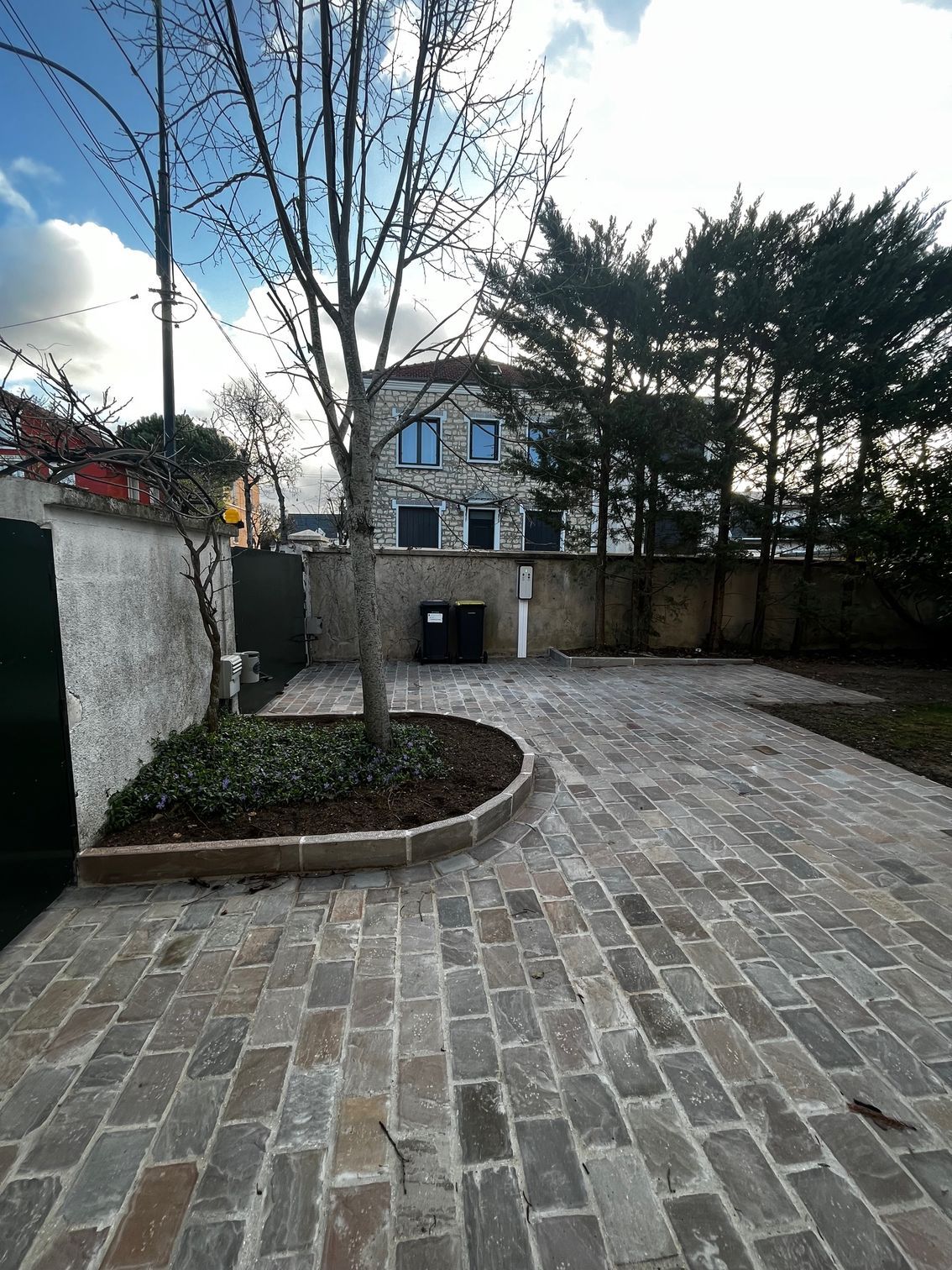 Cour pavée avec un petit parterre d'arbre, de hauts murs et une maison en arrière-plan sous un ciel nuageux
