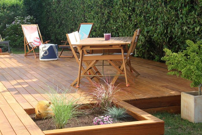 Terrasse en bois avec table, chaises, plantes en pot et parterre de fleurs surélevé dans un jardin.