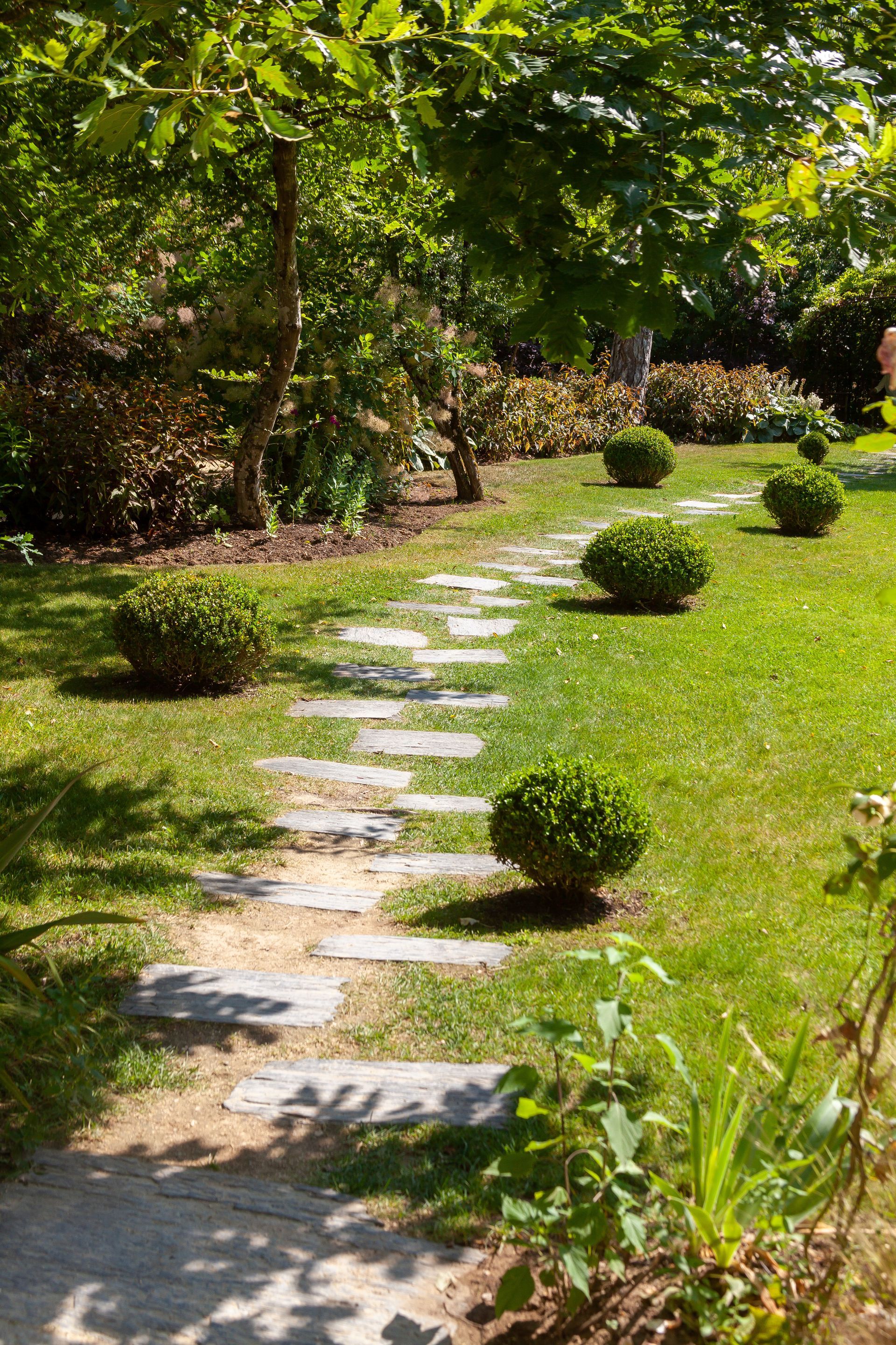 Un chemin de dalles traverse un jardin avec de l'herbe, des arbustes et des arbres.