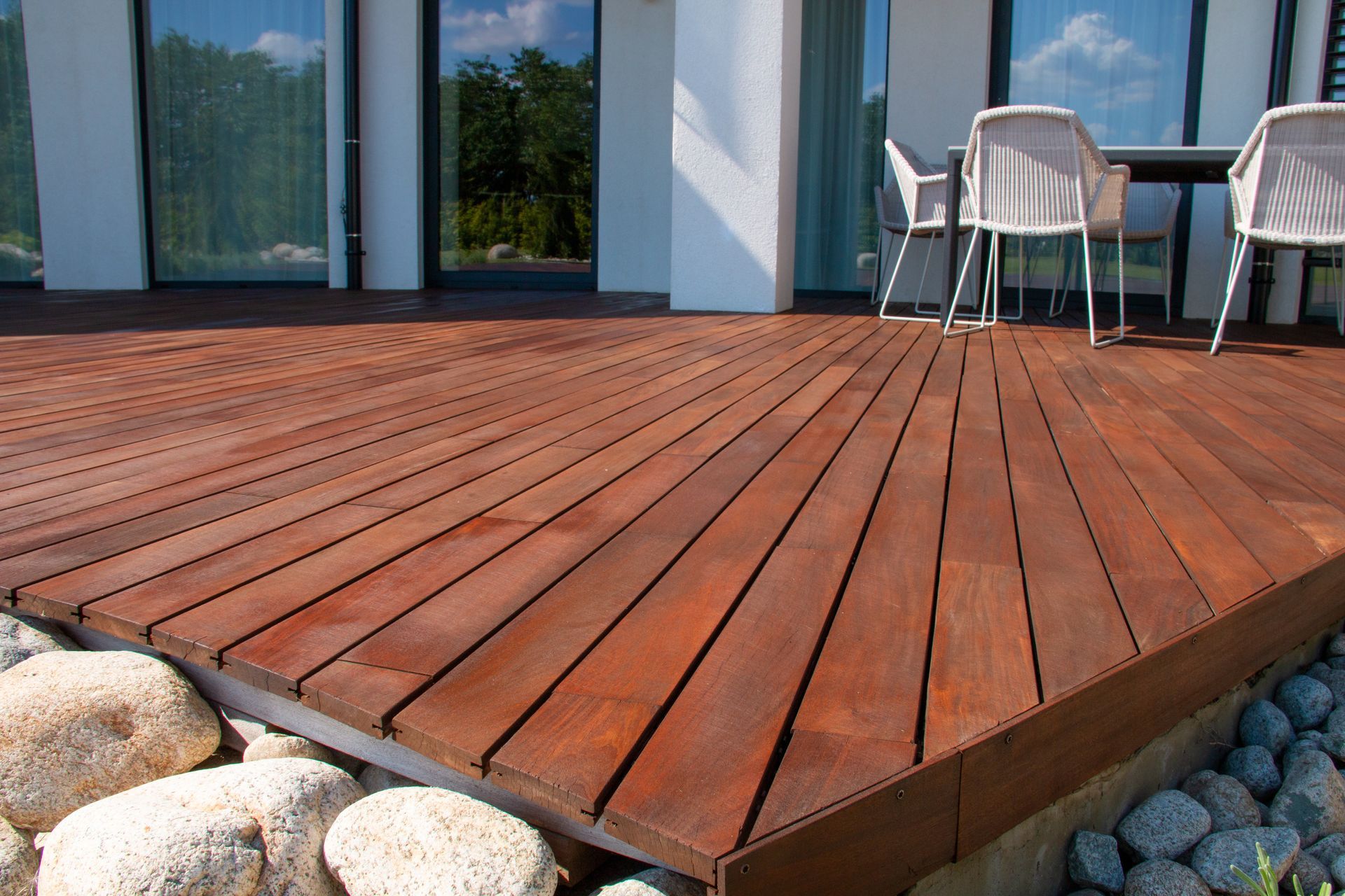 Terrasse en bois attenante aux portes vitrées, avec chaises et rochers devant.