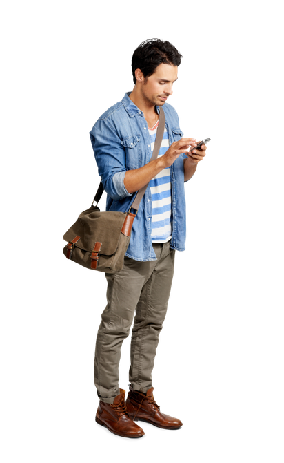 Un homme debout, regardant son téléphone, porte une chemise en jean, un pantalon gris, des bottes et une sacoche.