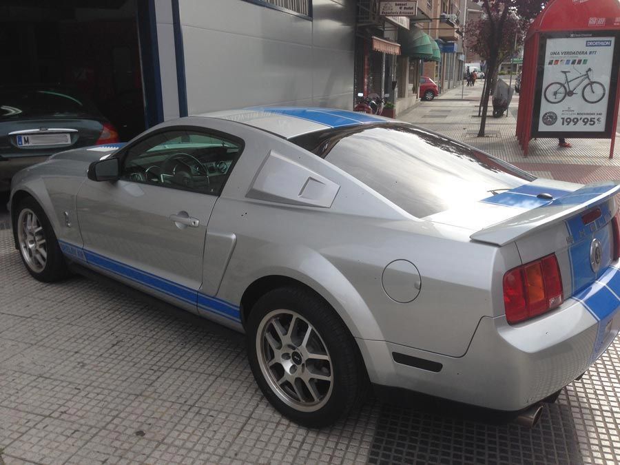 Un Mustang plateado y azul está estacionado frente a una parada de autobús.
