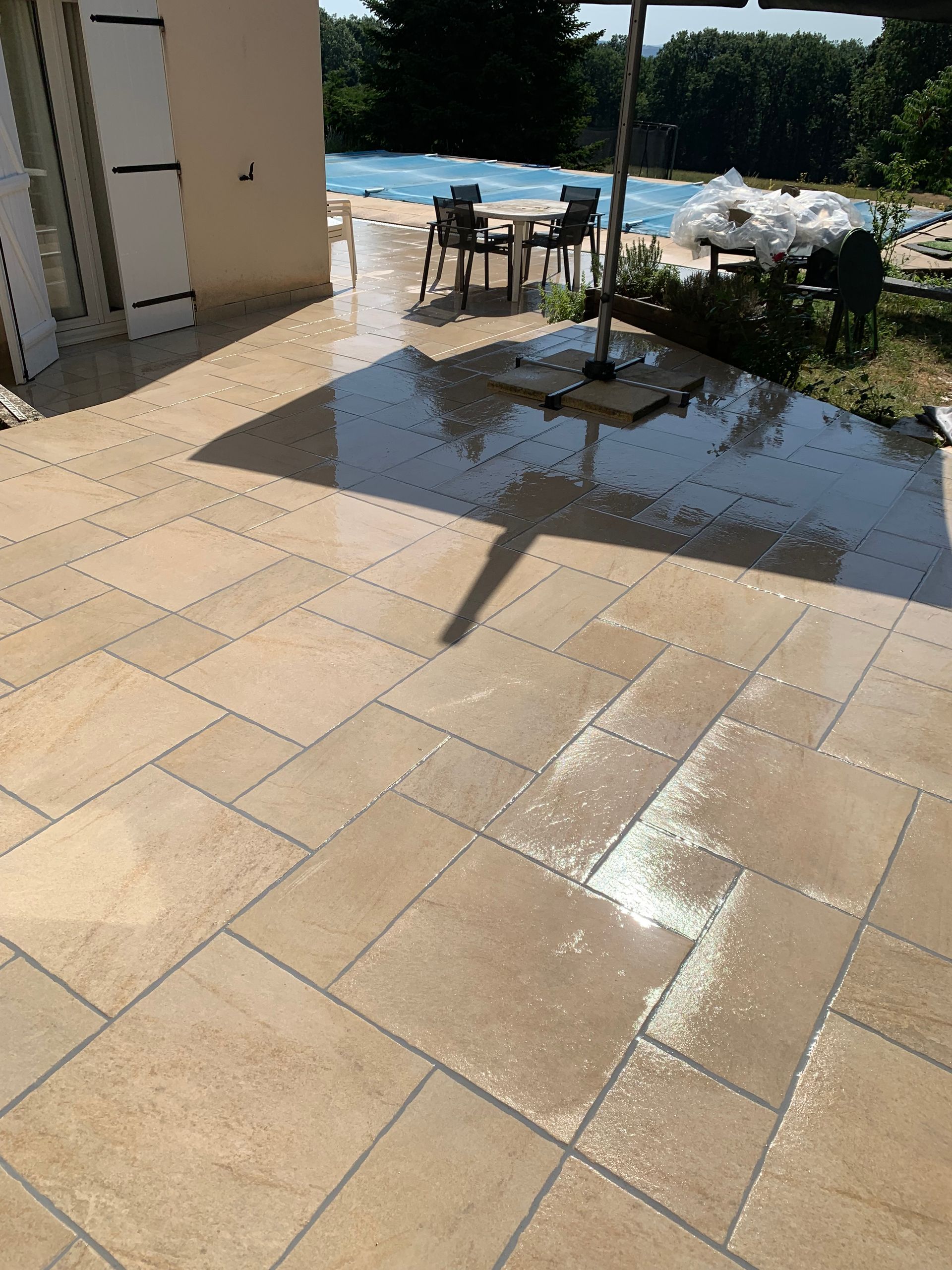 Patio ensoleillé avec carrelage beige, zones humides, une table, des chaises et une piscine en arrière-plan