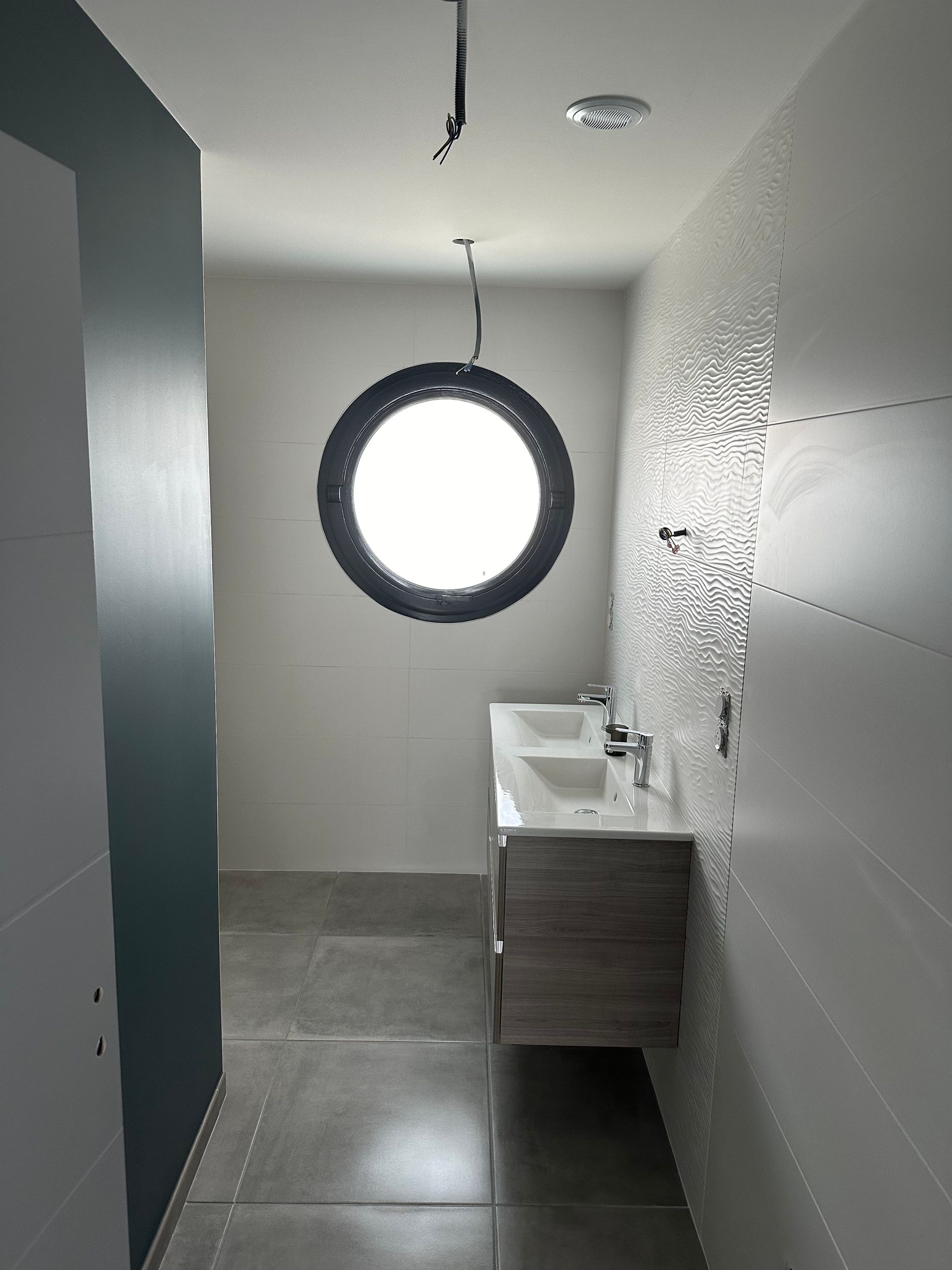 Toilettes modernes étroites avec fenêtre ronde, sol gris, lavabo blanc et cloison de cabine bleu foncé