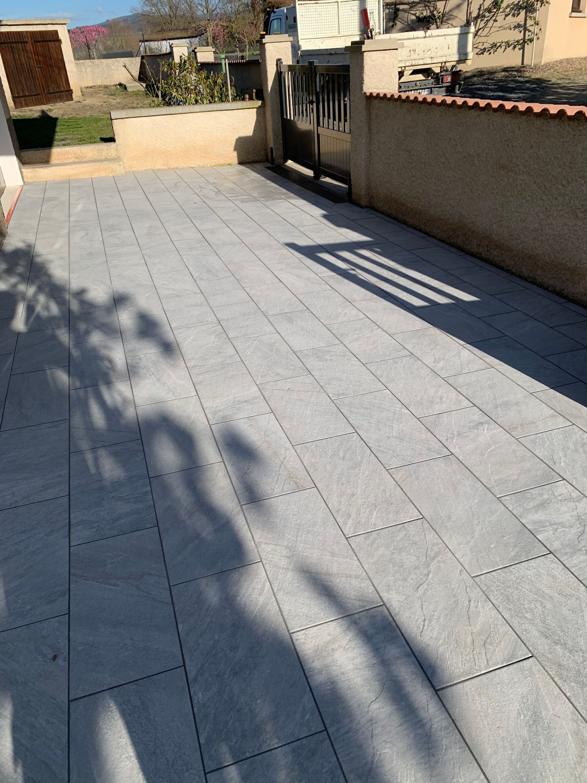 Terrasse carrelée et ensoleillée, adossée à un mur en stuc, avec de longues ombres et des collines lointaines au-delà d'une clôture.
