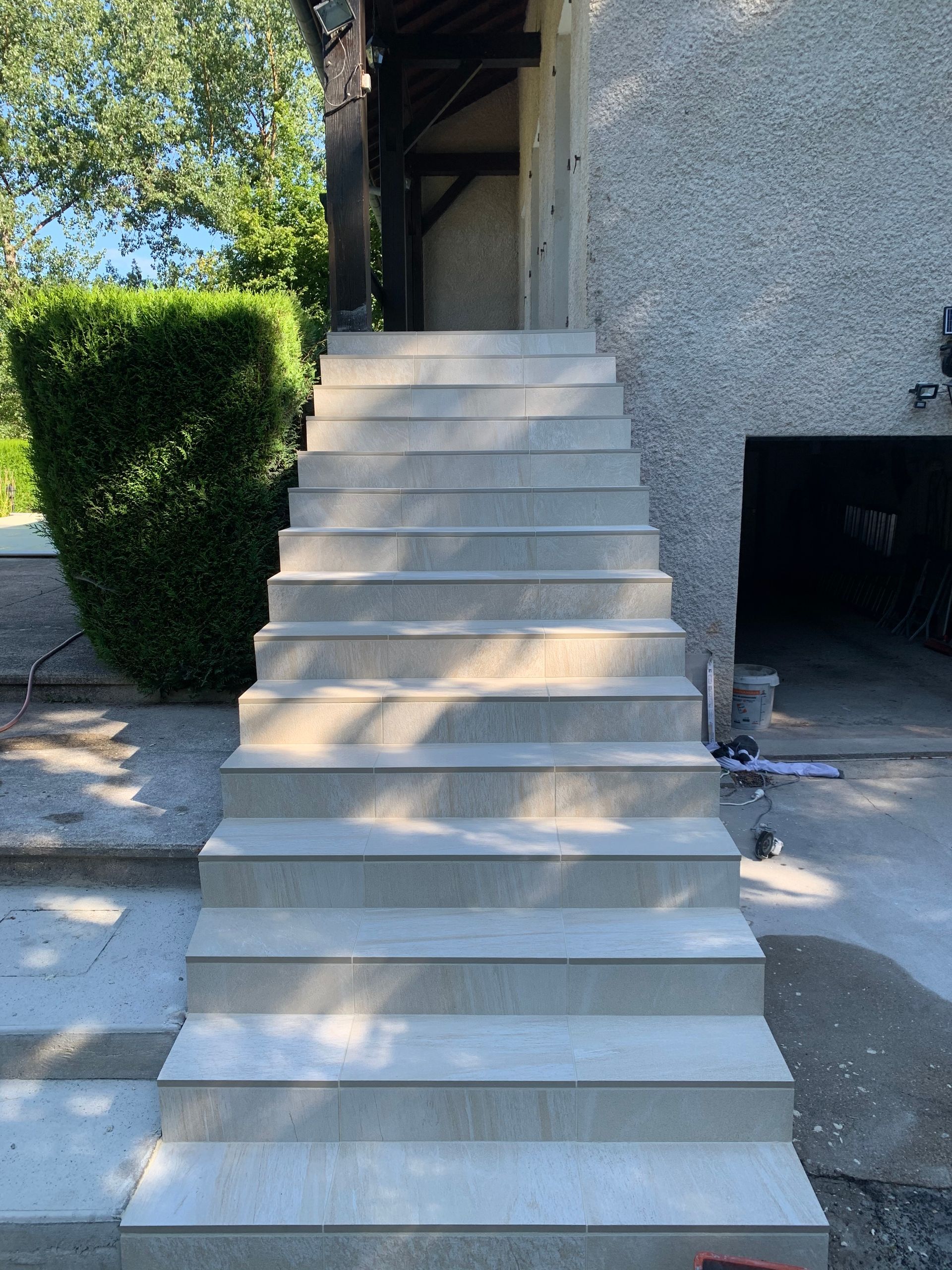 Un escalier en béton mène à l'entrée d'un bâtiment, bordé d'une haie et d'un mur gris.