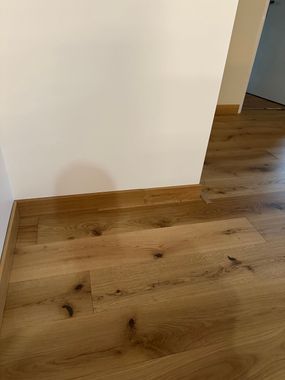 Un sol en bois jouxte un mur blanc, avec une porte ouverte donnant sur une pièce en arrière-plan.