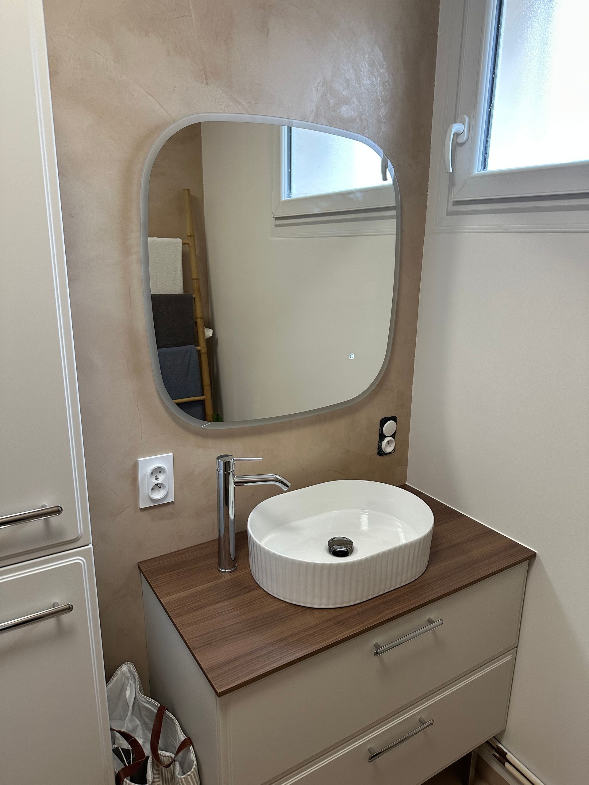 Petit meuble-lavabo de salle de bain avec lavabo rond, miroir et murs beiges sous une fenêtre.