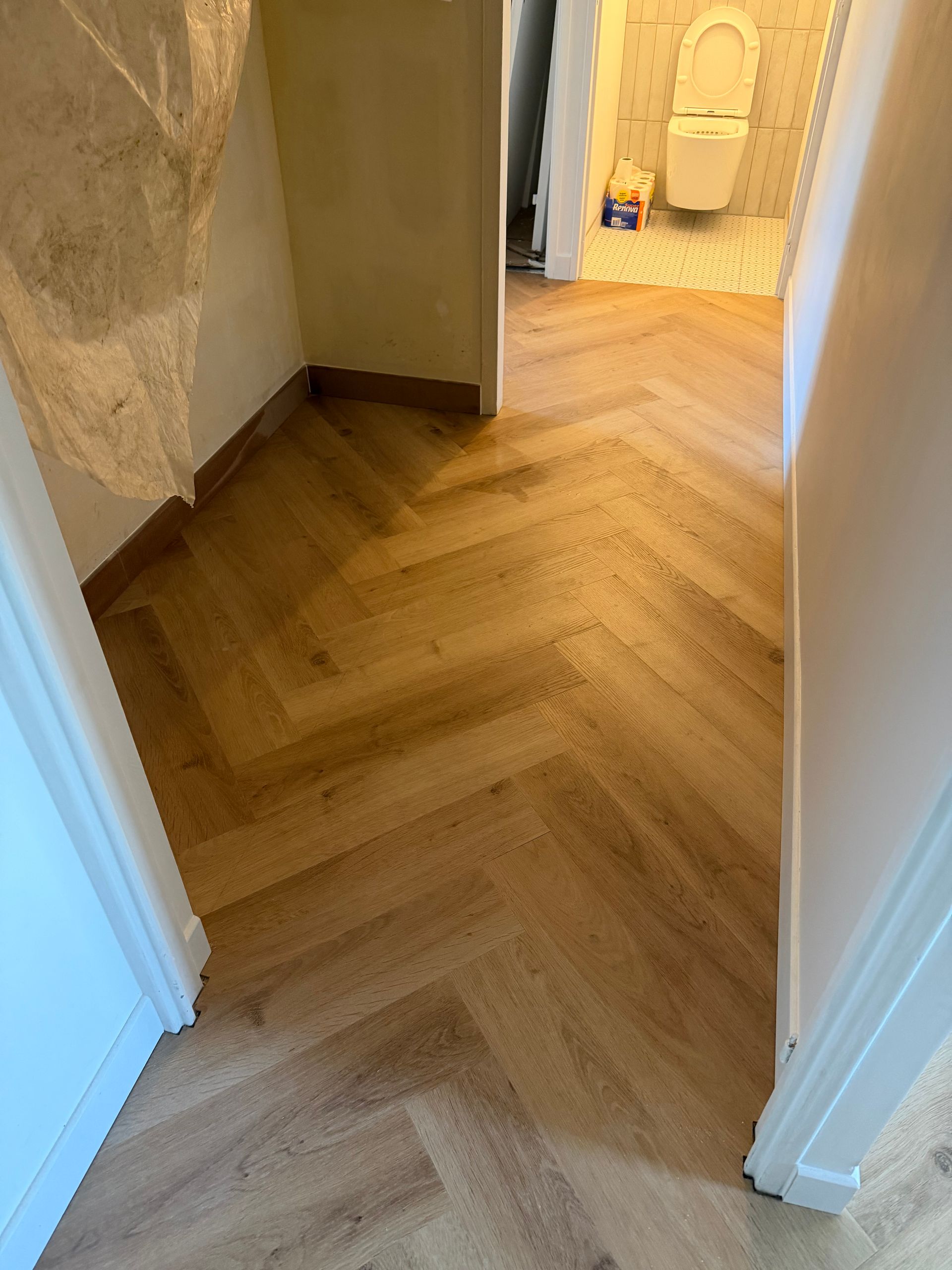 Couloir avec parquet à chevrons menant à une porte de salle de bains éclairée.