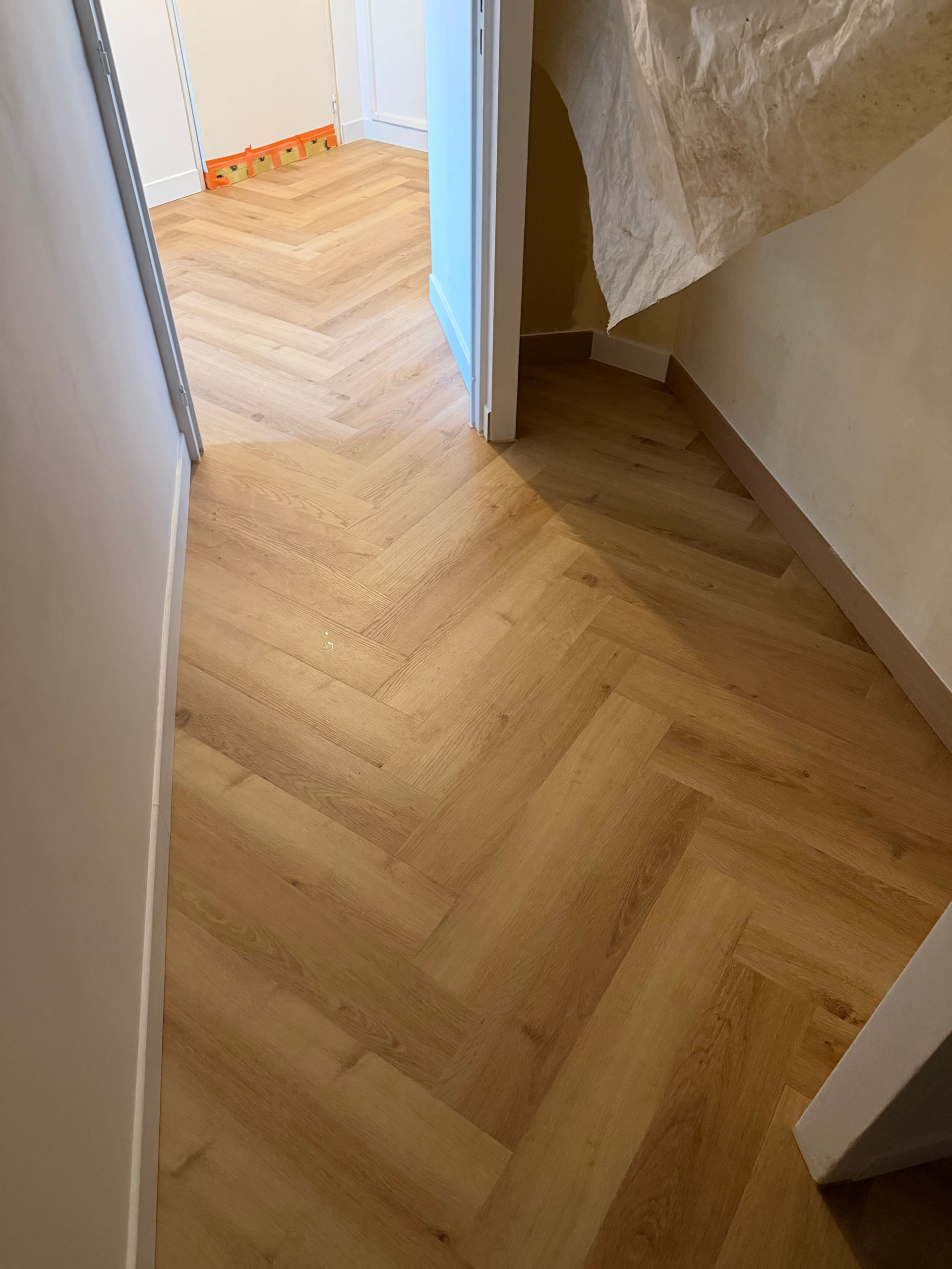 Couloir étroit avec parquet clair à chevrons, murs blancs et petit palier d'escalier près d'une porte.