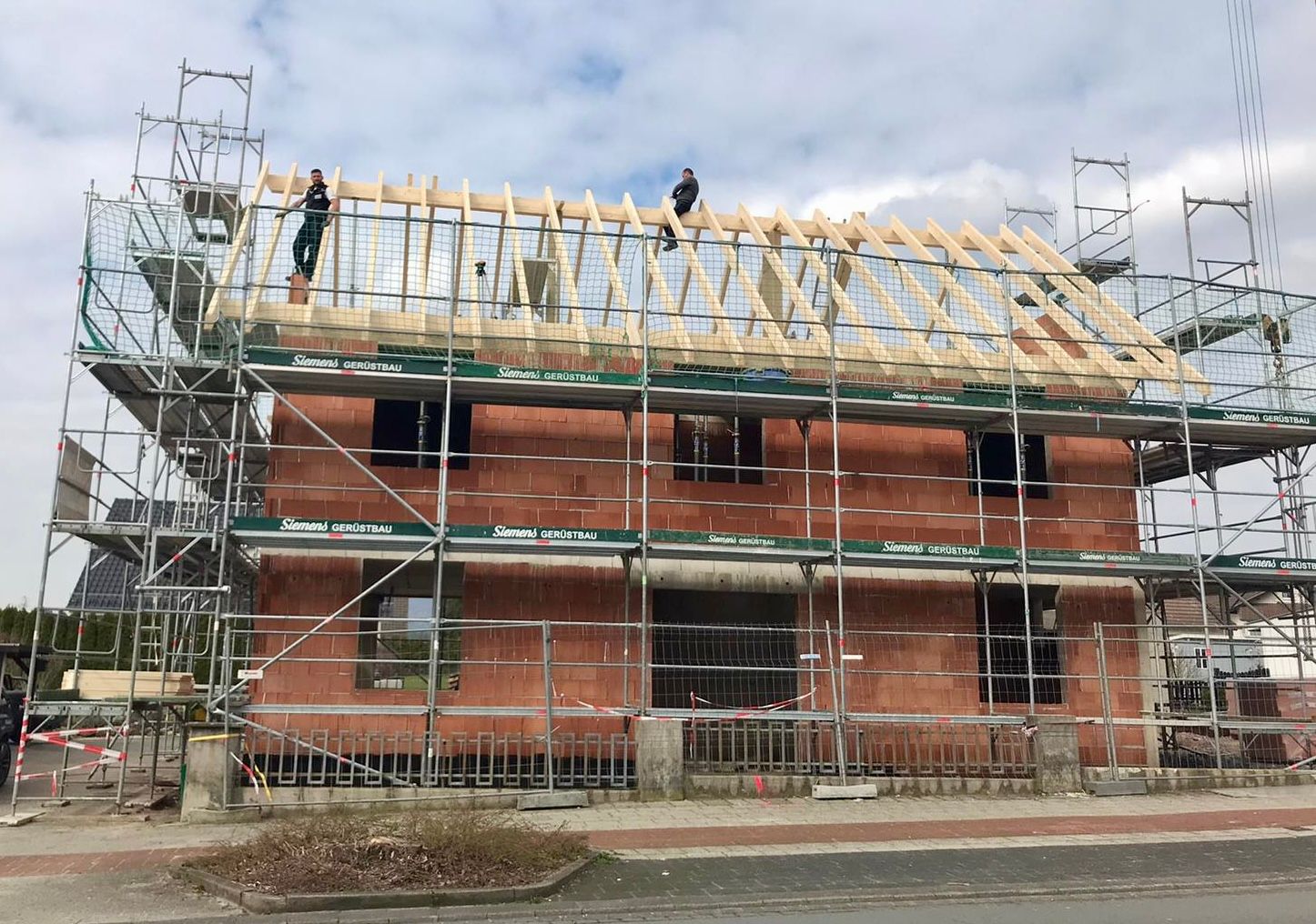 Bauarbeiter errichten unter bewölktem Himmel das Holzdach eines roten Backsteinhauses, das von einem Gerüst umgeben ist.