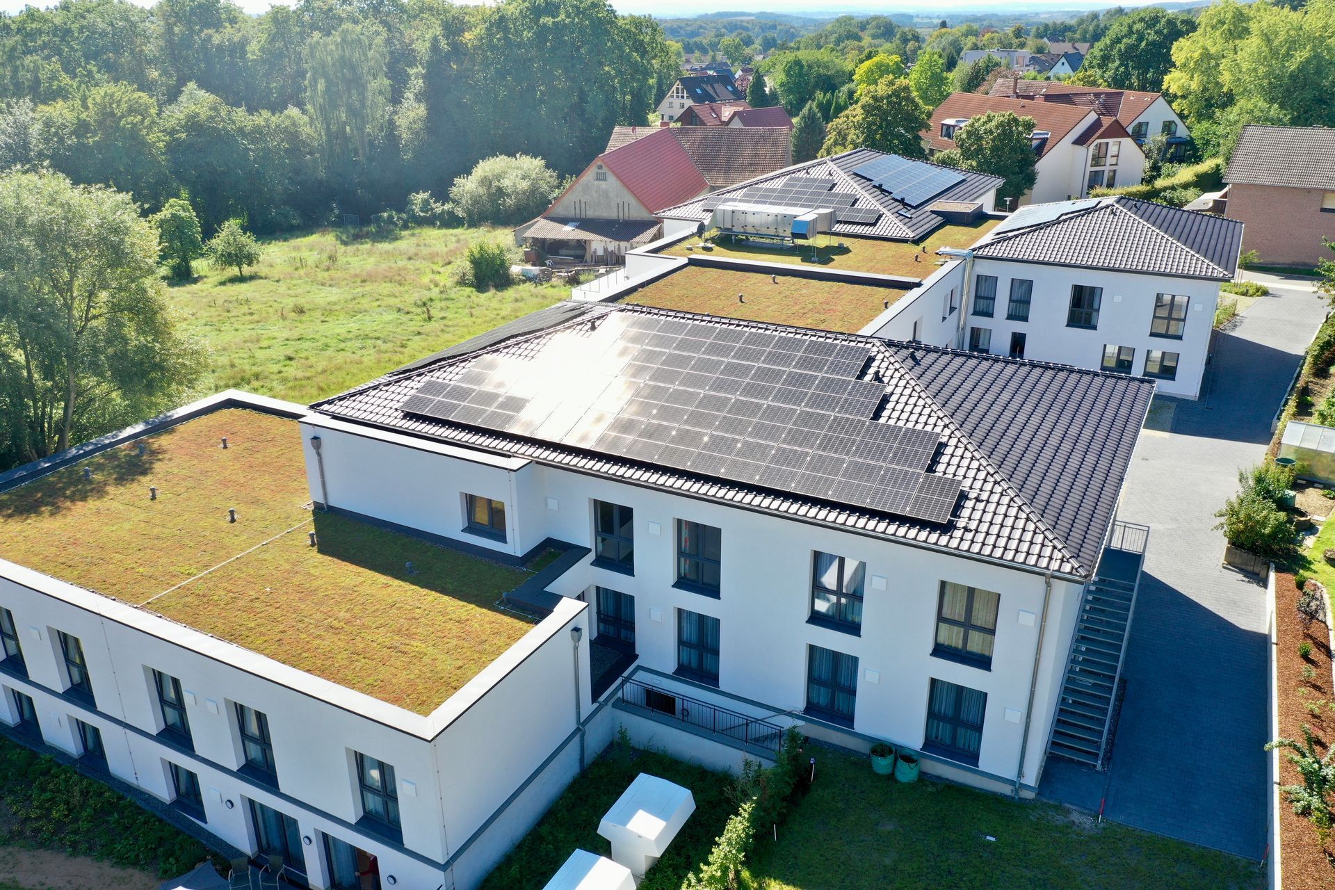 Luftaufnahme eines weißen, mehrstöckigen Gebäudes mit Gründach und Solaranlage an einem sonnigen Tag.