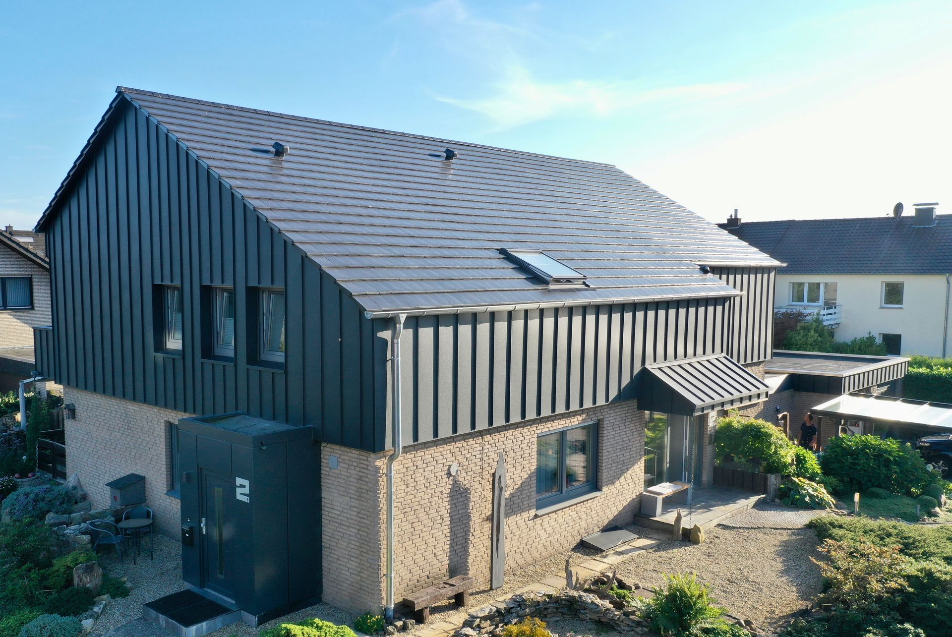 Ein zweistöckiges Haus mit beigefarbenem Backsteinsockel, dunkelgrauer vertikaler Fassadenverkleidung und Metalldach