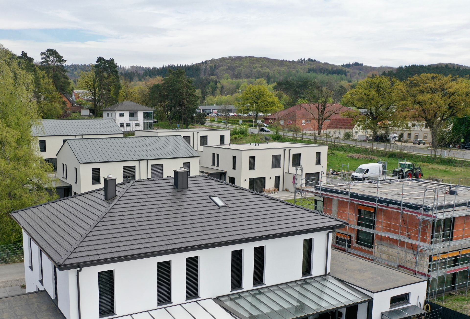 Luftaufnahme von modernen weißen Häusern mit grauen Ziegeldächern in einer Vorstadtlage, im Hintergrund Bäume und Hügel.