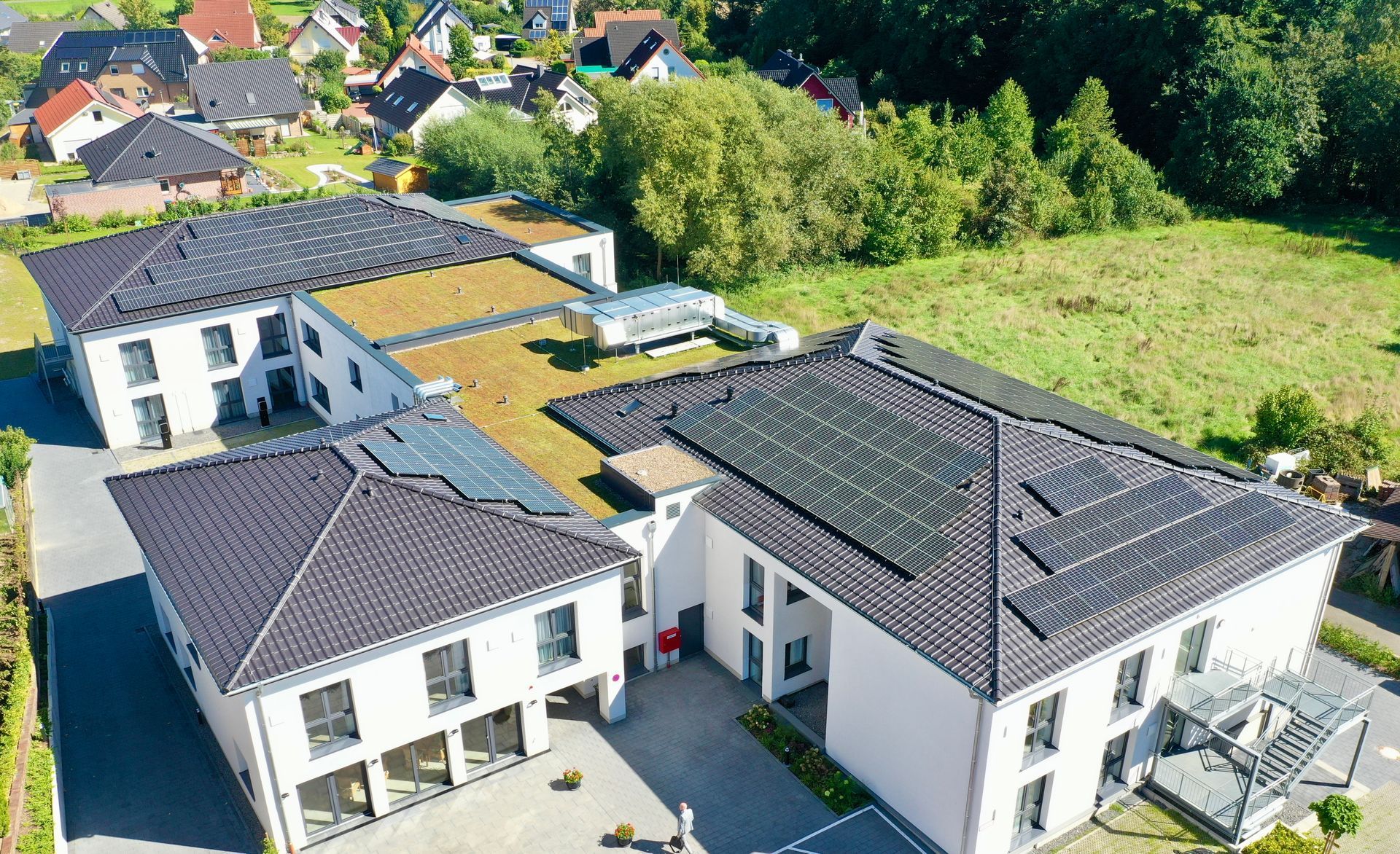 Luftaufnahme eines weißen, mehrflügeligen modernen Gebäudes mit dunklen Ziegeldächern und Solarpaneelen