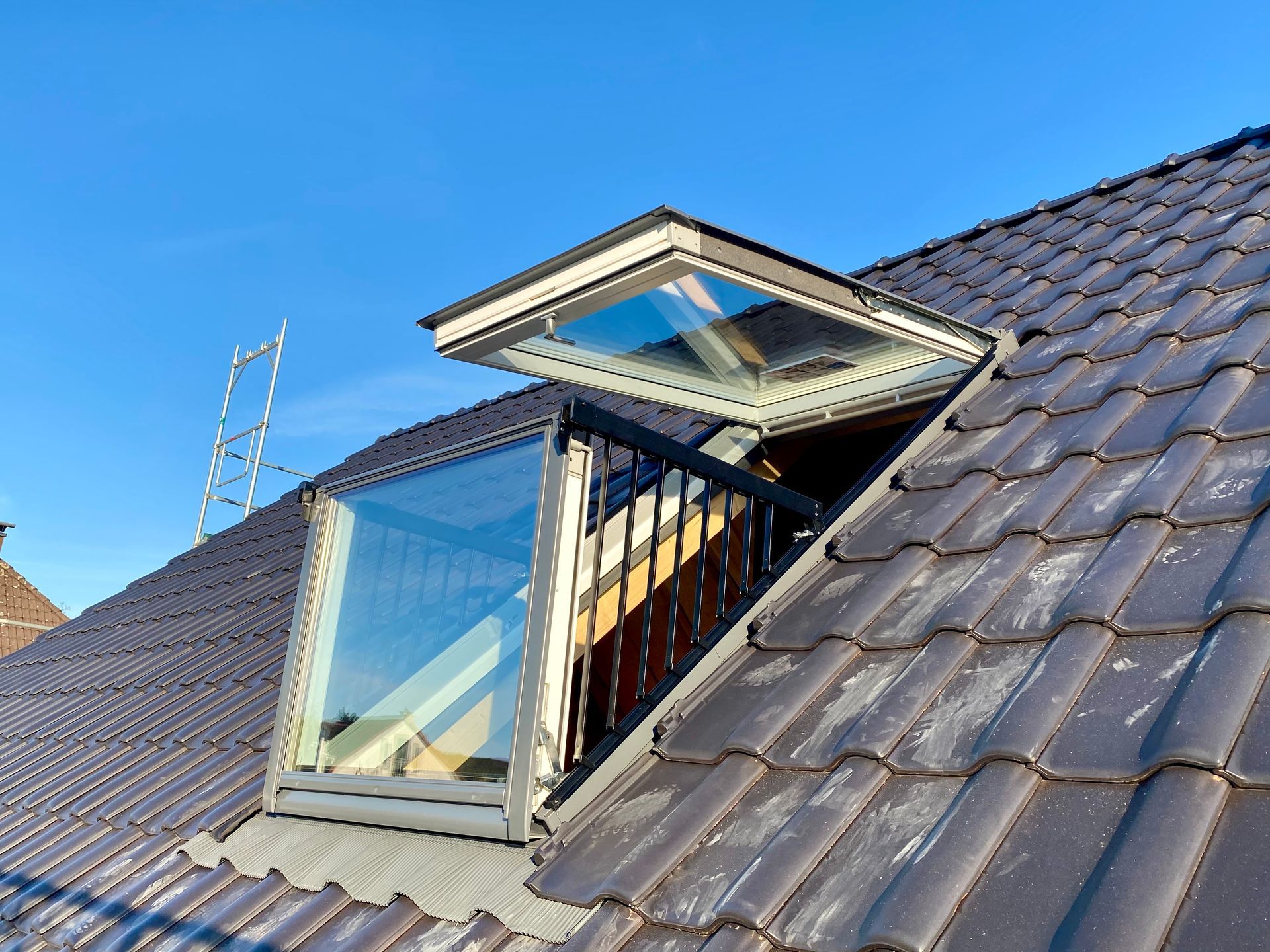 Ein vollständig geöffnetes Balkonfenster auf einem dunklen Ziegeldach vor strahlend blauem Himmel.